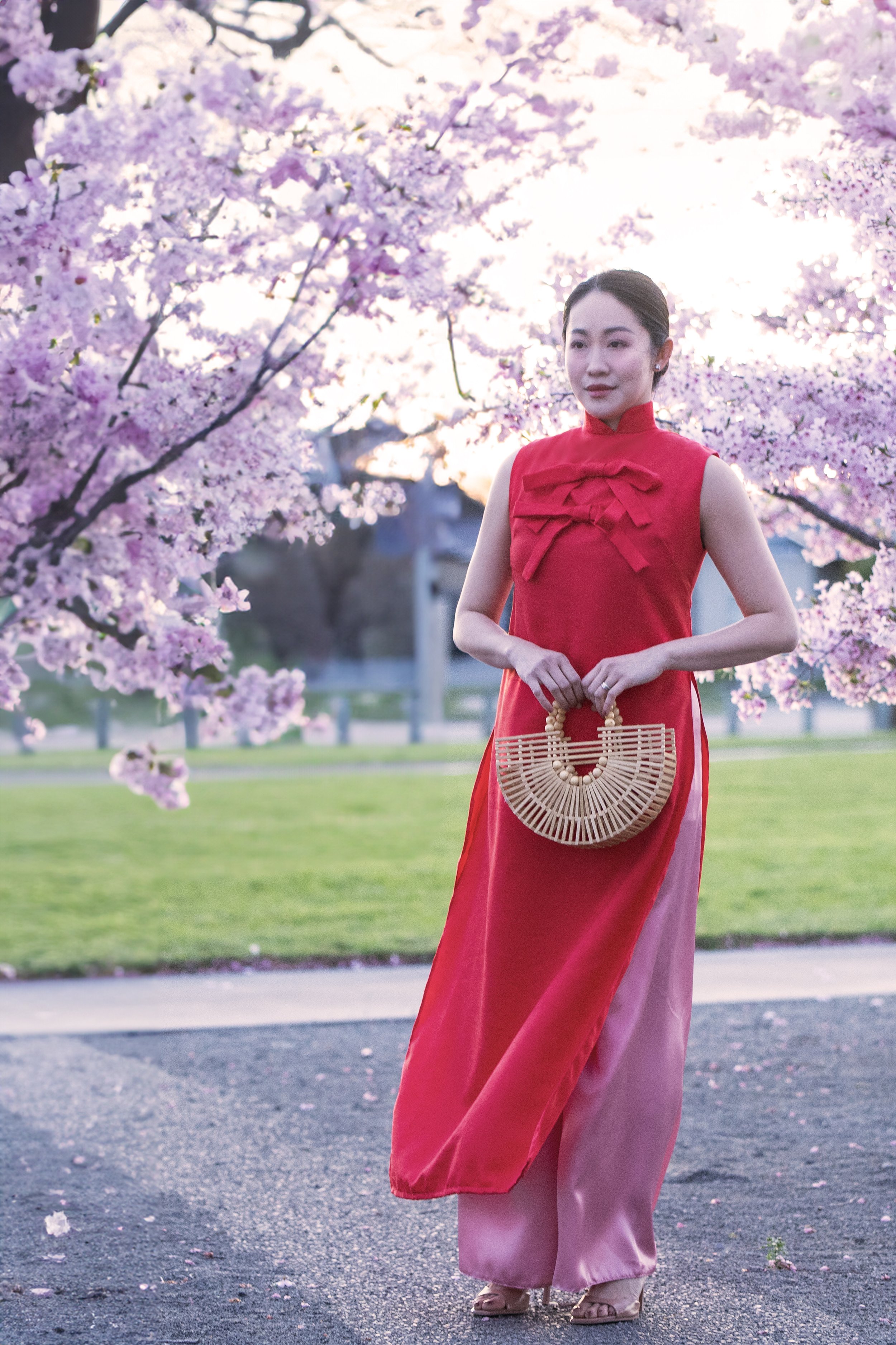 A woman in a traditional red and pink Vietnamese dress holding a bamboo handbag, standing near pink cherry blossom trees with a park in the background.