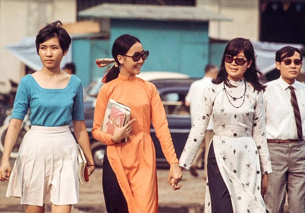 Four women and one man walking on the street, some holding magazines, dressed in vintage clothing and sunglasses, with retro cars in the background.