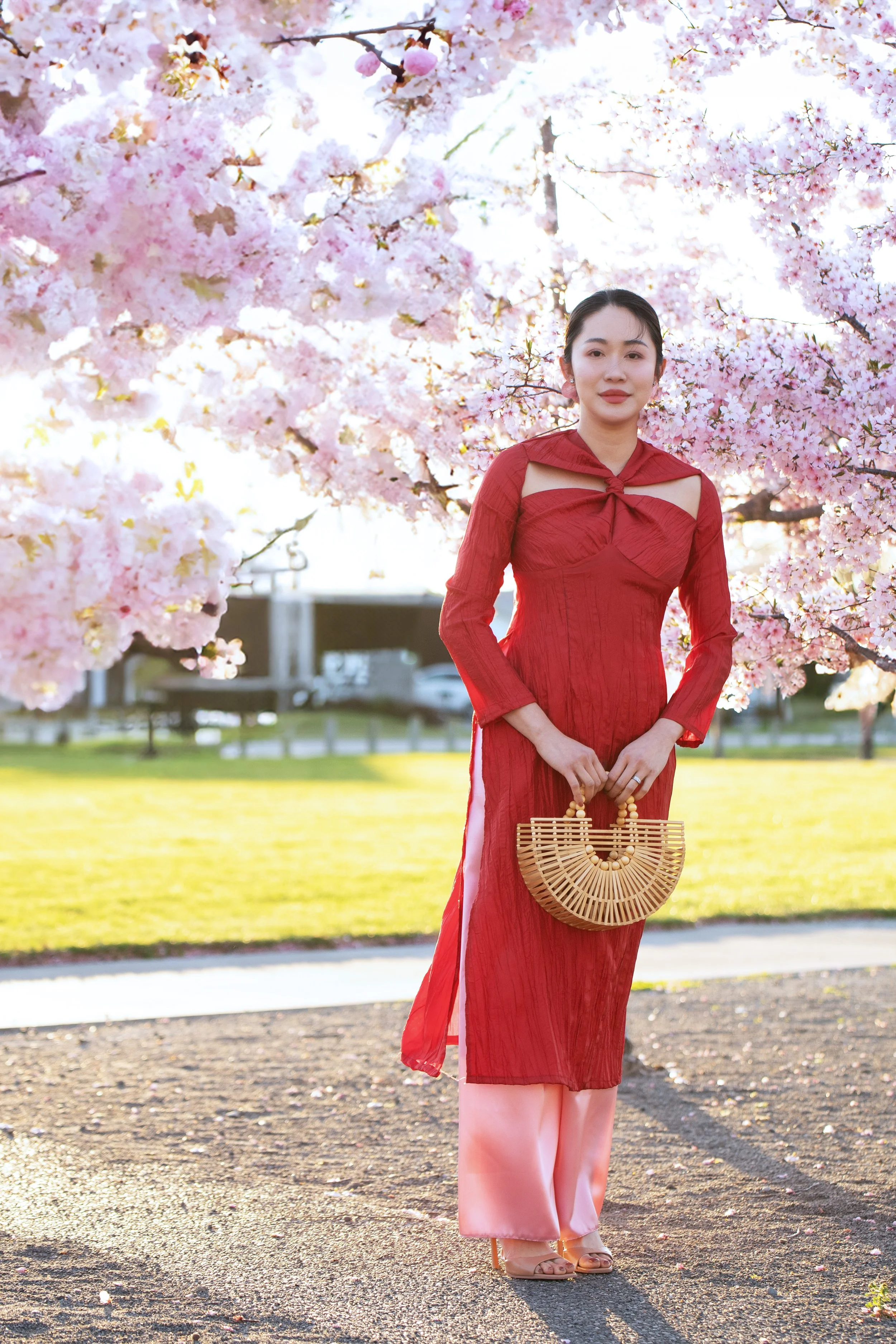 A woman in a red dress and pink pants holding a bamboo handbag standing outdoors under cherry blossom trees with pink flowers.
