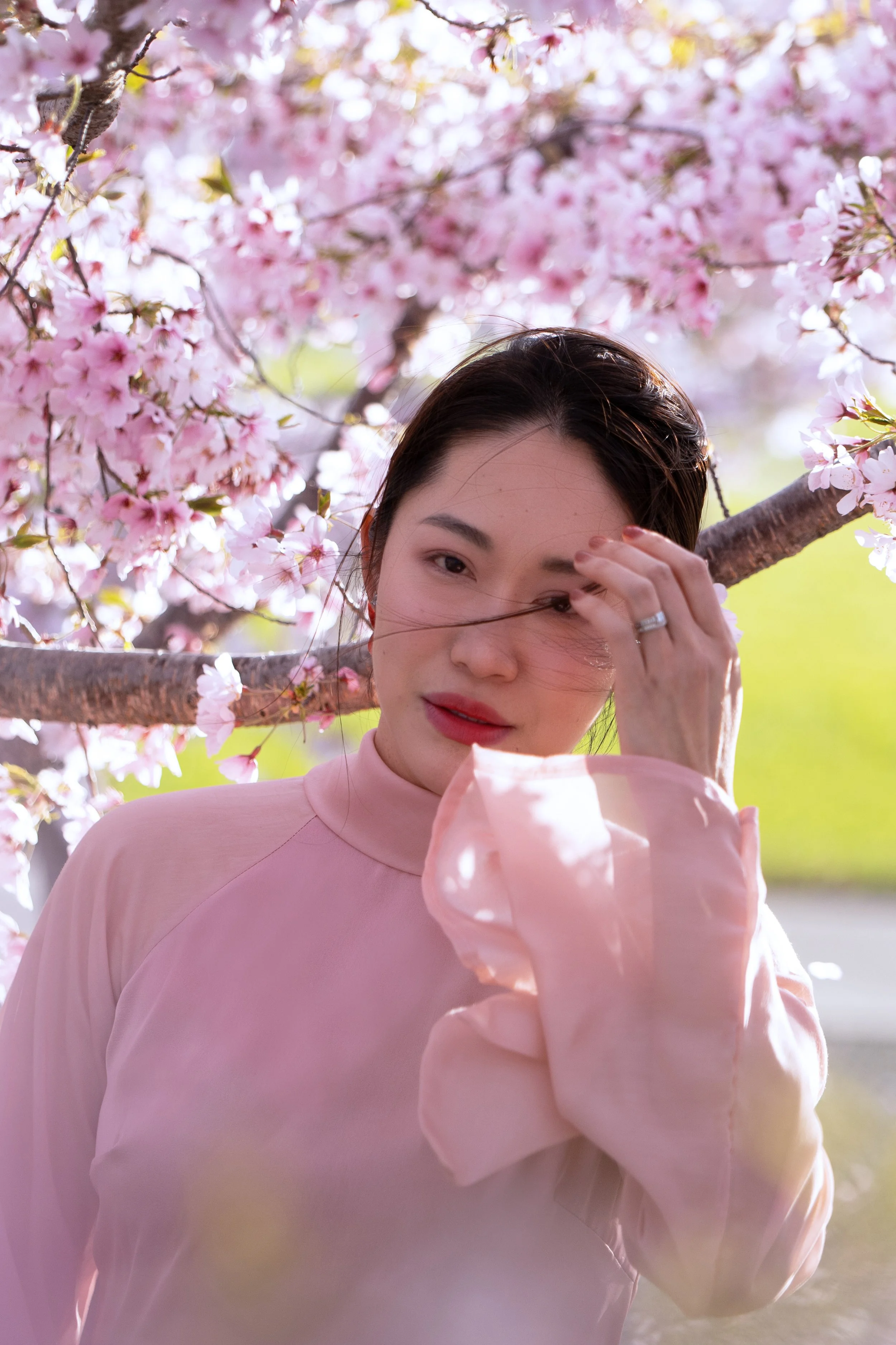 A woman with dark hair standing among pink cherry blossoms, holding her hand to her face and looking into the camera.
