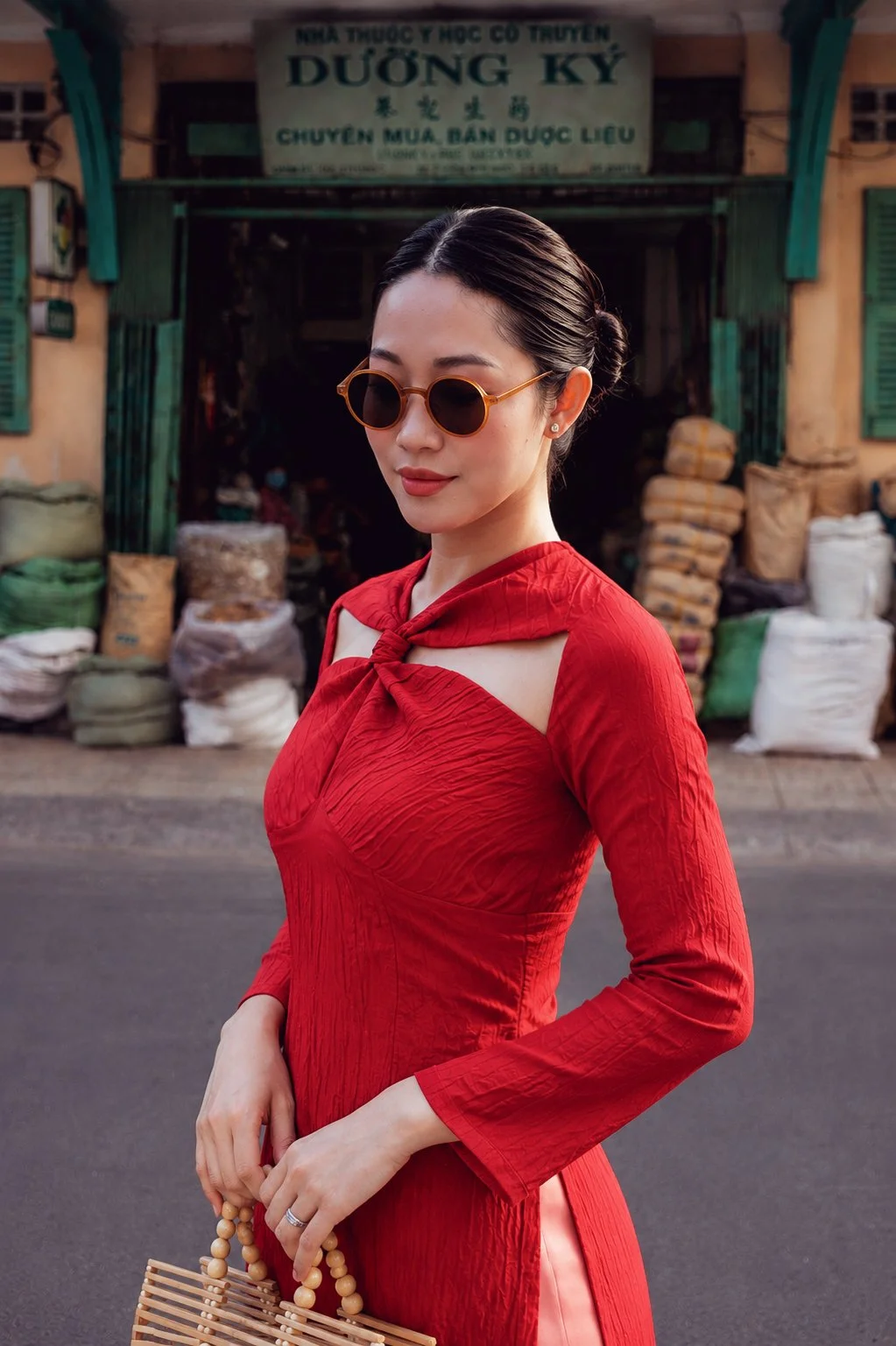 A woman wearing a red dress and sunglasses standing outside in front of a shop with stacks of bags or sacks. She is holding a wooden handbag and has her hair styled in a bun.