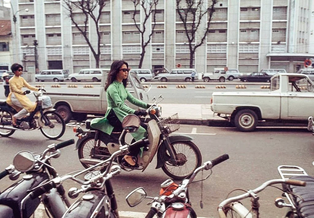 A woman in green riding a scooter on city street, with bicycles and cars nearby, and tall buildings in the background.