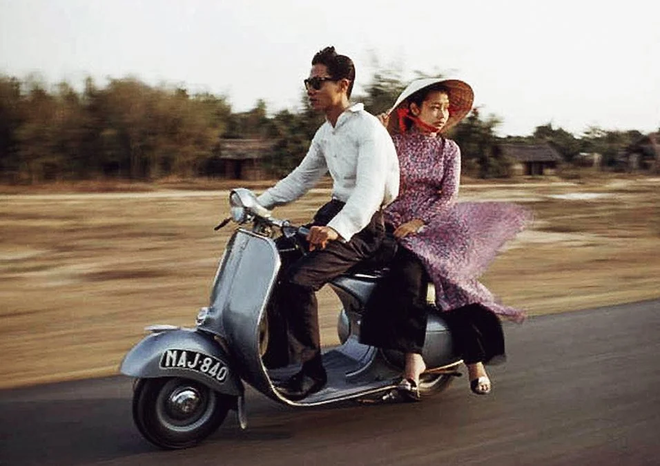 A man and woman riding a motorcycle on a road in a rural area; the woman is wearing a large hat and a flowing dress.