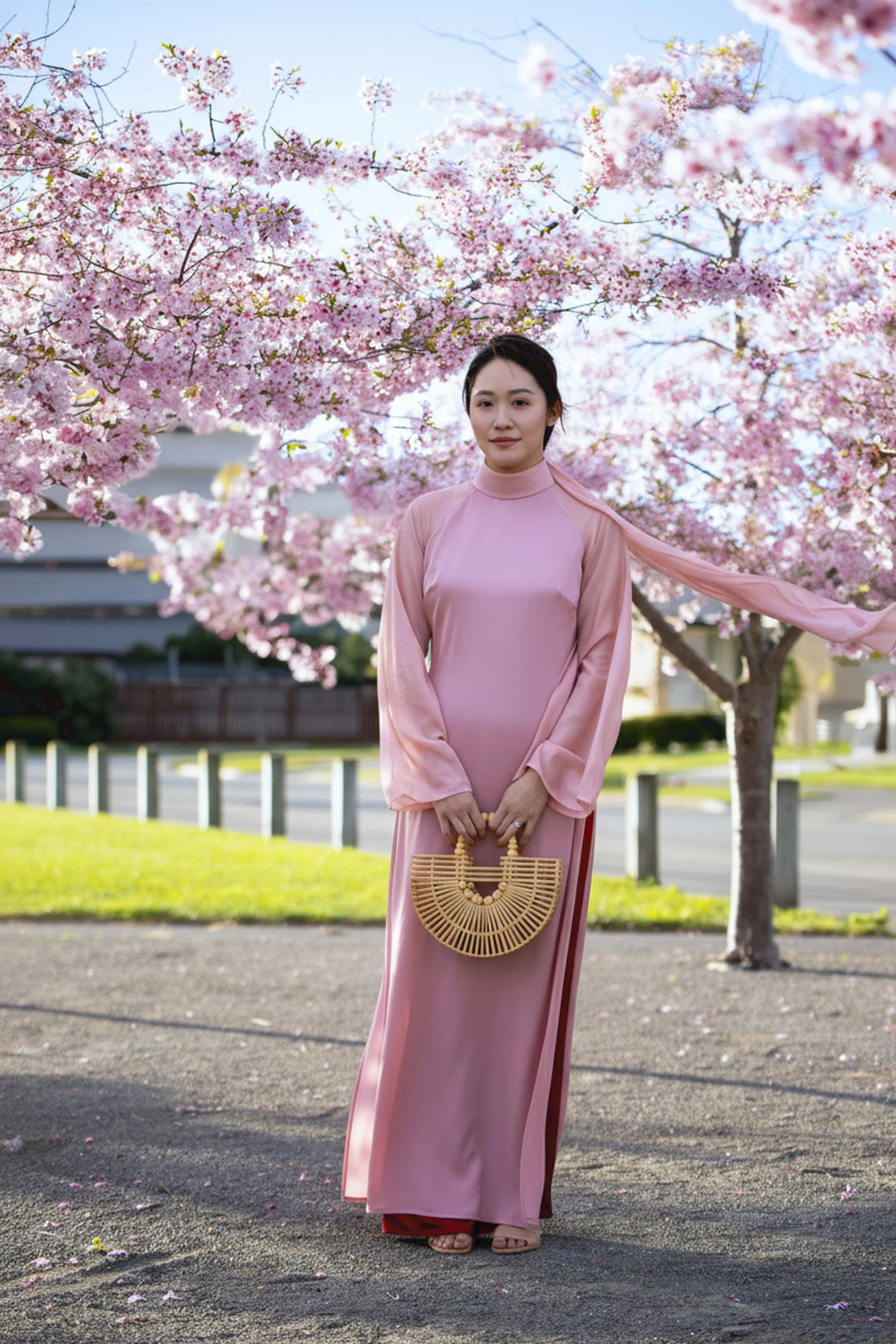 A woman in a pink dress holding a bamboo purse, standing outdoors in front of blooming cherry blossom trees on a sunny day.