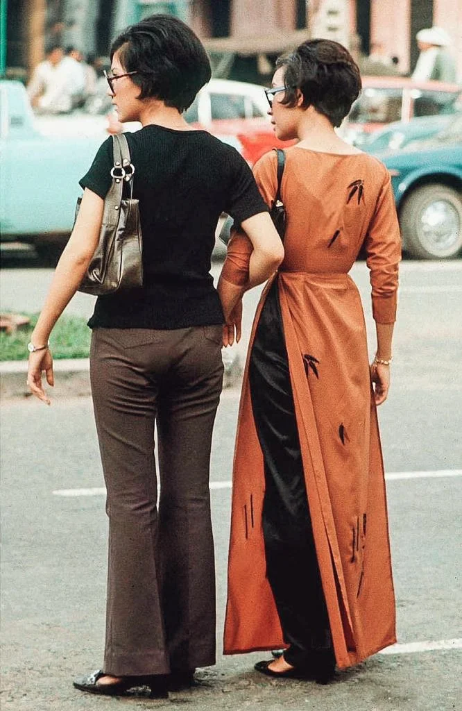 Two women with short dark hair and glasses walk together on a city street during the daytime. One woman is dressed in a black top and brown pants, carrying a shiny gray handbag, while the other is wearing an orange traditional dress with black accent