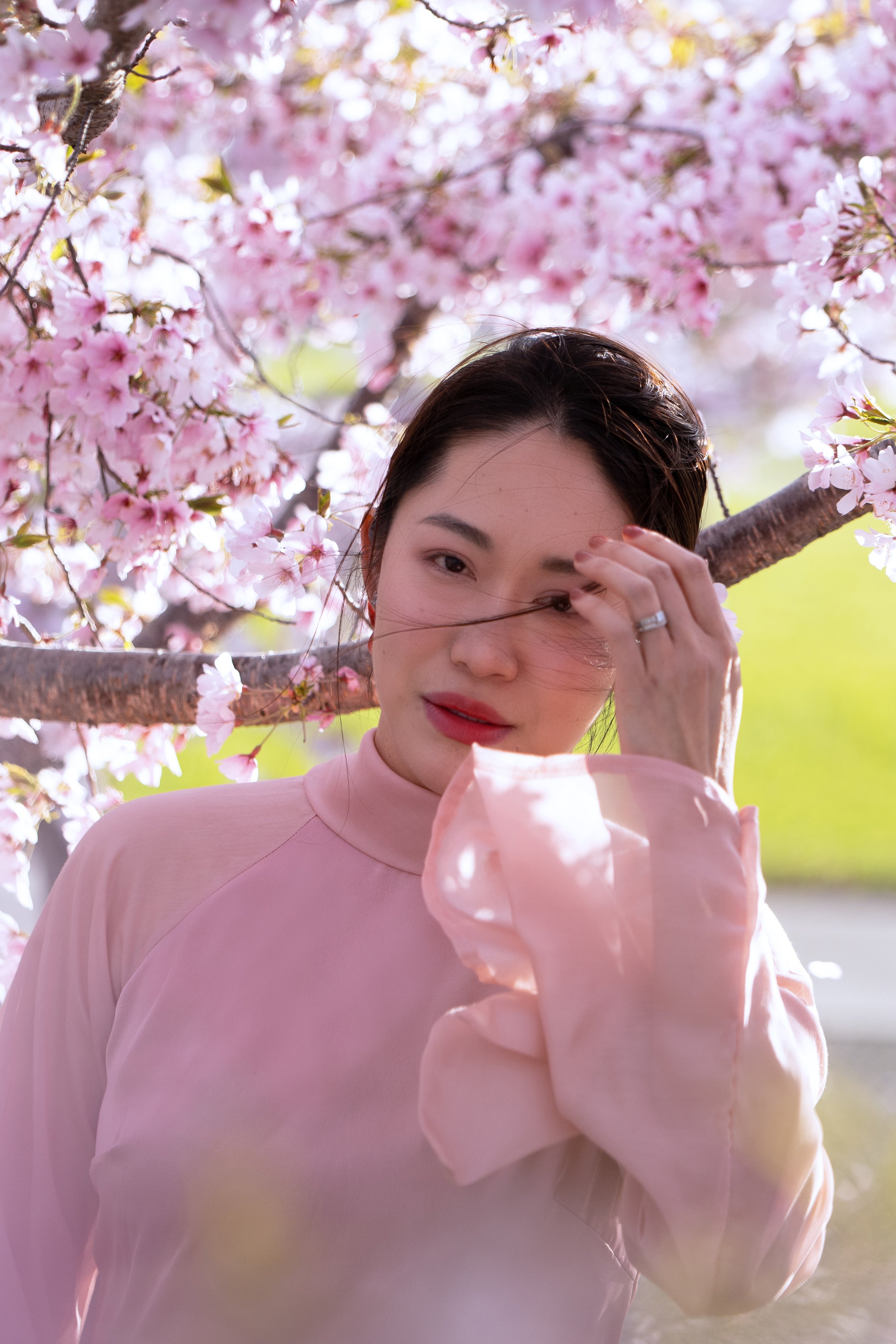A woman with dark hair, wearing a pink top, standing amidst pink cherry blossoms, adjusting her hair with one hand, outdoors on a sunny day.