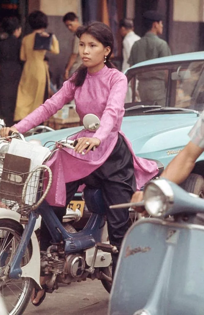 A young woman in a pink traditional dress riding a bicycle on a busy street with vintage vehicles and people in the background.