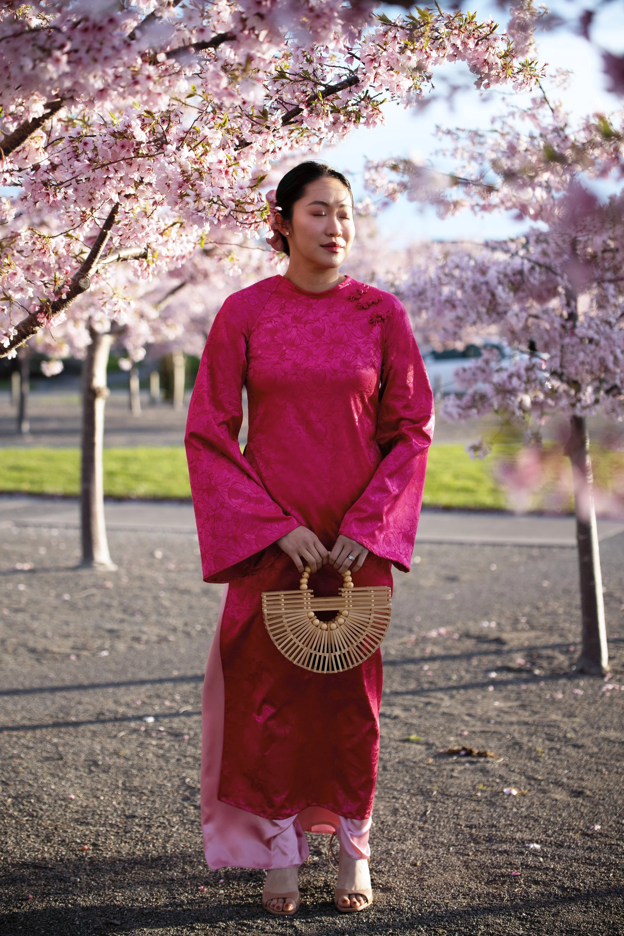 A woman wearing a bright pink traditional Asian dress standing among blooming pink cherry blossom trees.