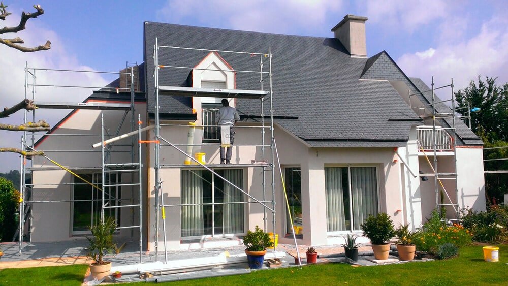 Maison en rénovation avec échafaudage et un ouvrier peintre sur le toit.