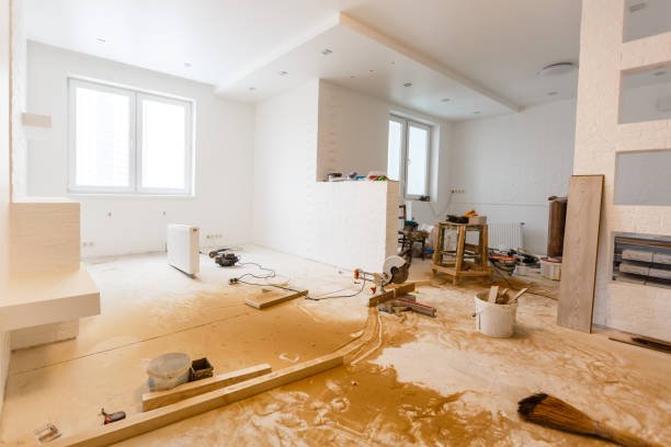 Pièce en rénovation avec sol couvert de poussière, outils et matériaux dispersés. Murs blancs, grandes fenêtres, plafond avec spots intégrés. Échelle et radiateur visibles.