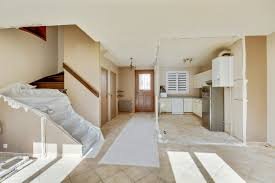 Intérieur d'une maison avec un escalier en bois, une cuisine moderne à droite et un sol carrelé beige.