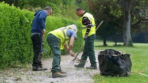Trois personnes portant des gilets de sécurité jaunes travaillent dans un jardin, utilisant des râteaux pour nettoyer un sentier en gravier. Un sac noir pour les déchets est visible à proximité.