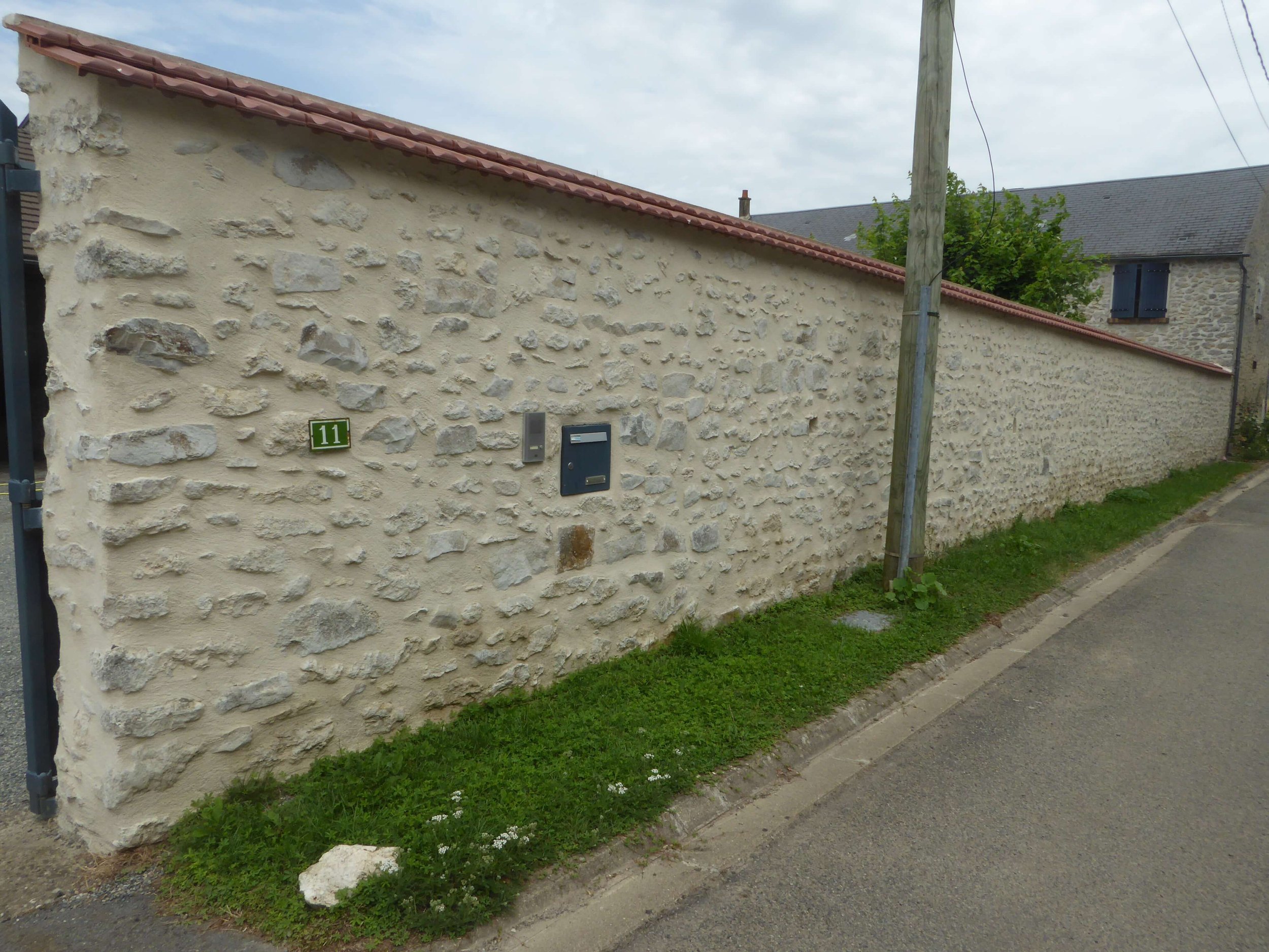 Un mur en pierre avec le numéro 11, une boîte aux lettres et une sonnette, longeant une rue pavée, avec un poteau téléphonique en bois à proximité.