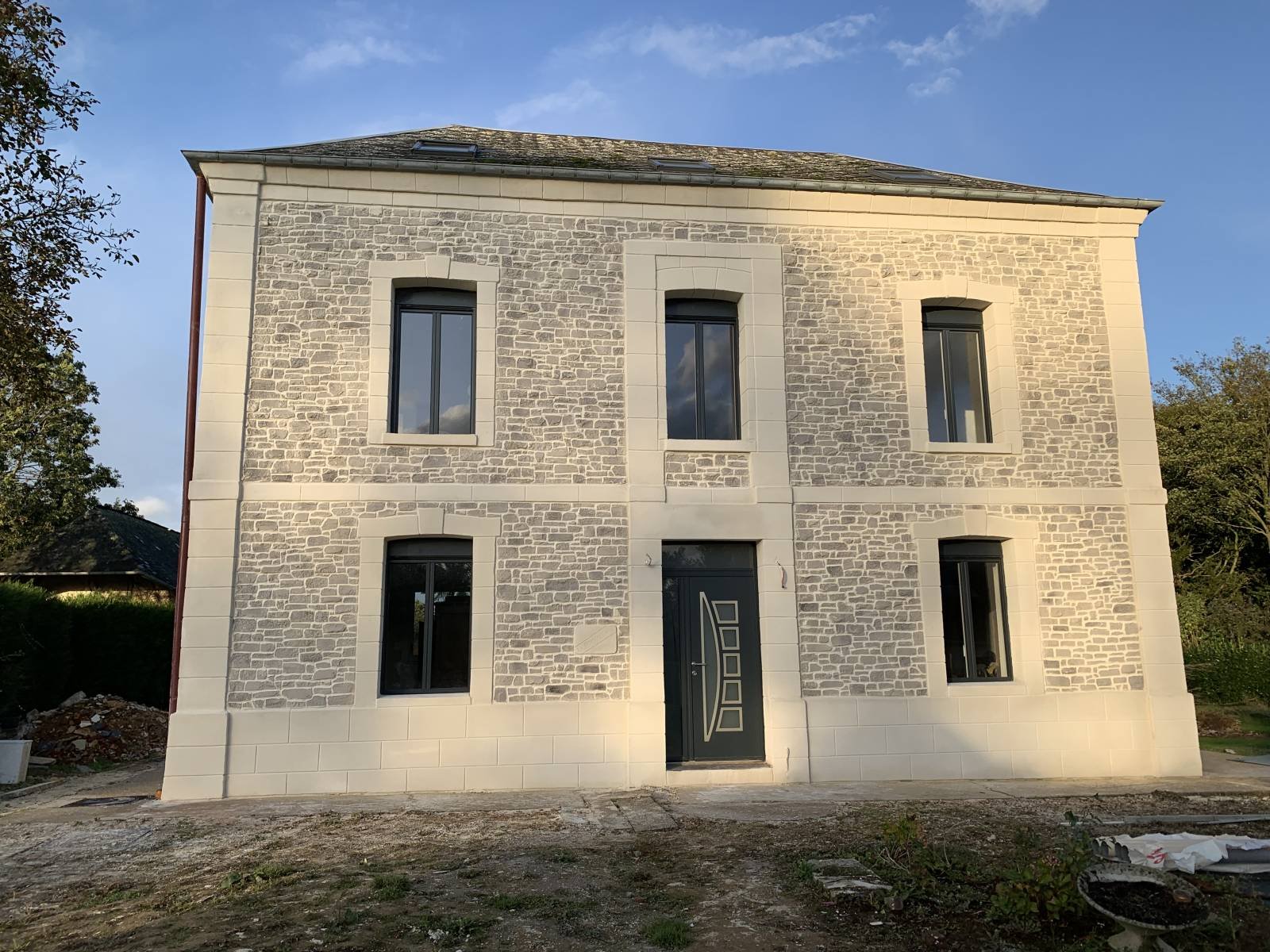 Maison en pierre à deux étages avec cinq fenêtres, une porte d'entrée moderne, et un toit incliné. Environnement extérieur naturel avec ciel bleu."