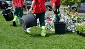 Trois jardiniers en uniforme rouge et vert portant des seaux noirs sur une pelouse, avec des fleurs et des plantes en arrière-plan.