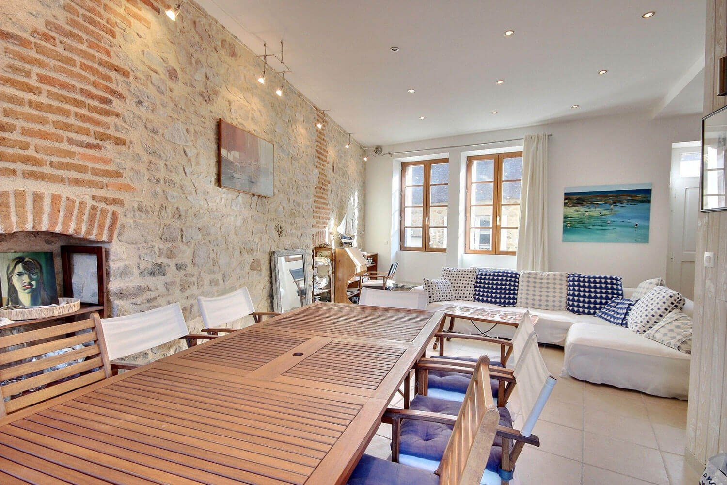 Salle de séjour avec mur en pierre, table en bois, chaises autour, canapé blanc avec coussins bleus, et fenêtres lumineuses.