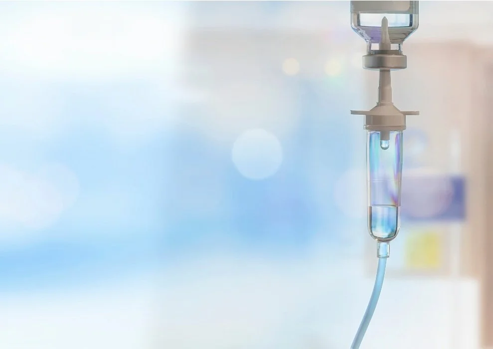 A medical IV drip hanging, filled with clear liquid, in a hospital setting.
