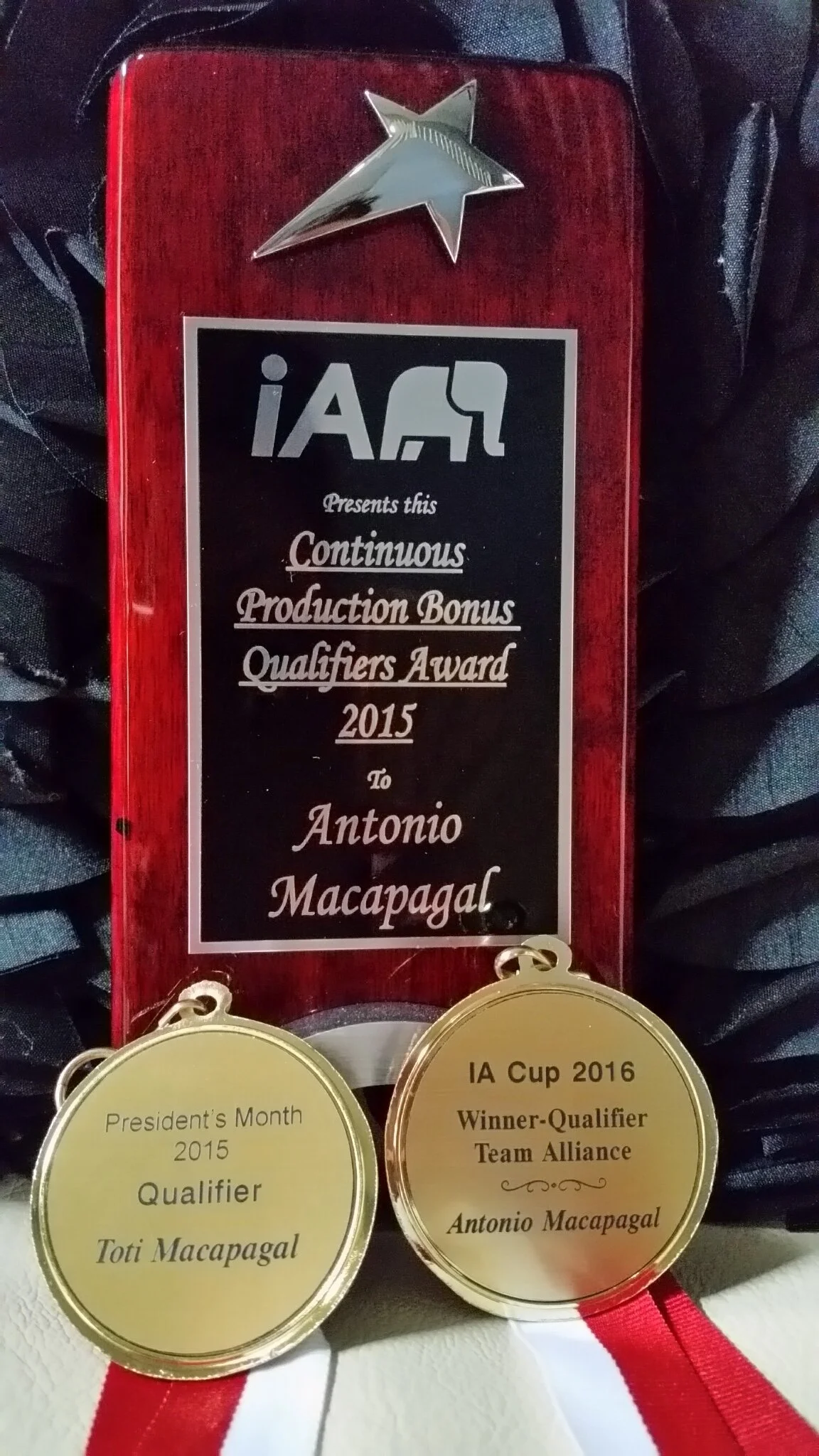 A wooden plaque with a metal star on top and two gold medals hanging in front of it. The plaque awards Antonio Macapagal a 2015 bonus qualification award, and the medals commemorate his qualification honors.