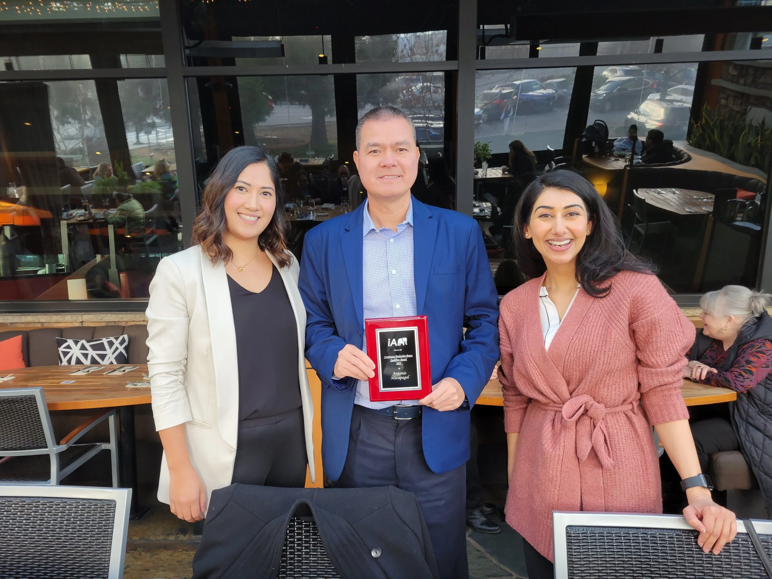 Three people standing together in a restaurant, the man in the middle holding a plaque, smiling at the camera.