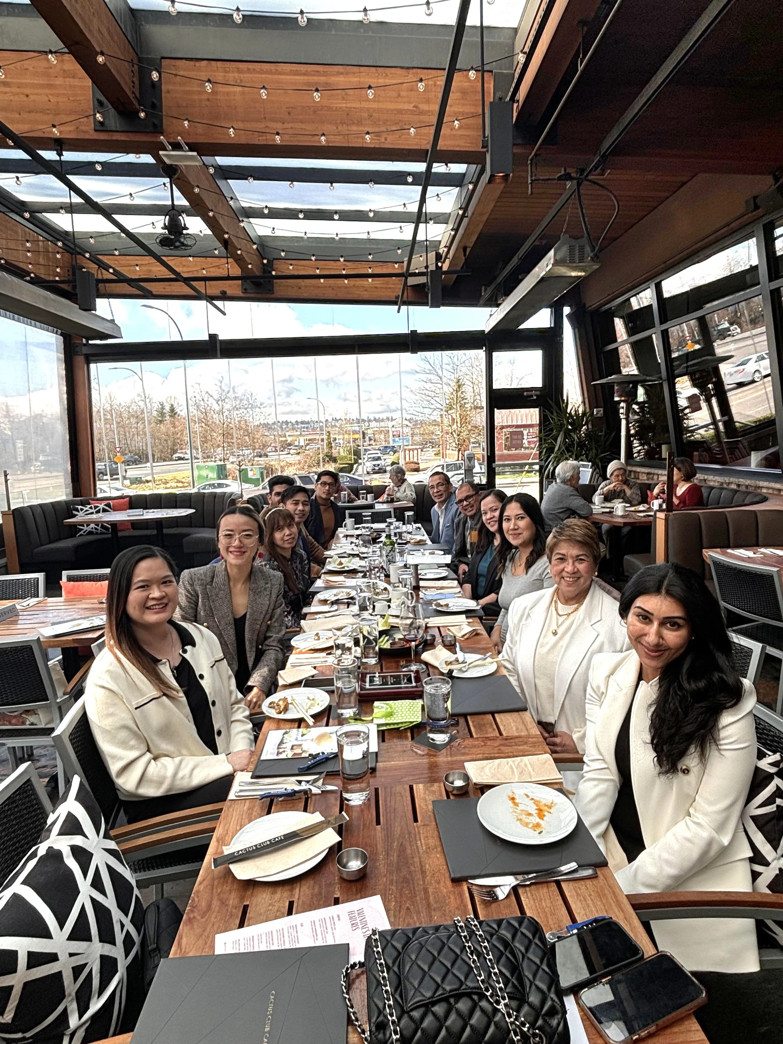 A group of people seated around a long dining table in a restaurant with large windows and wooden ceiling, enjoying a meal.