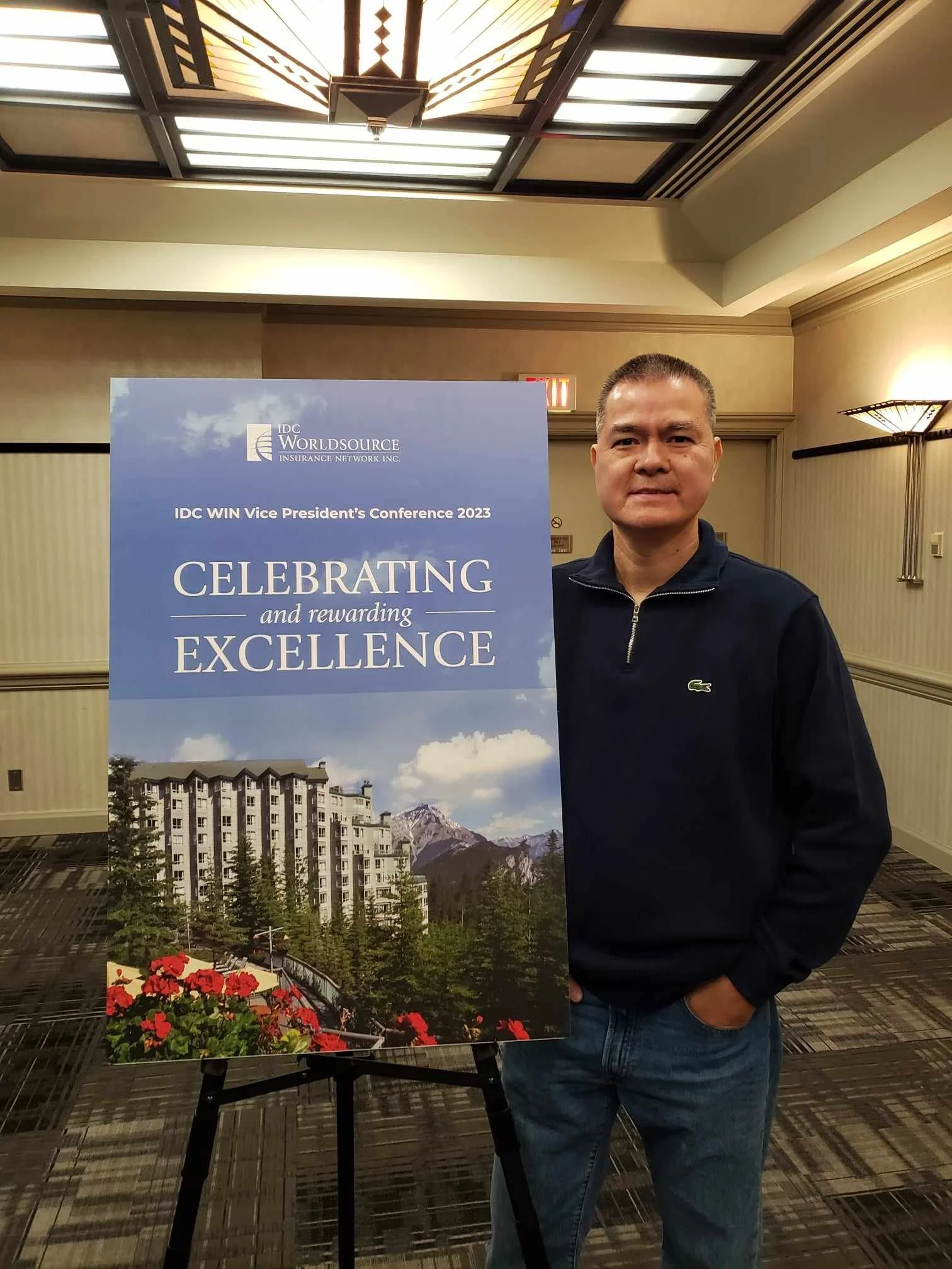 A man standing next to a sign displaying the text for the IDC WIN Vice President's Conference 2023, with a background image of buildings and mountains.