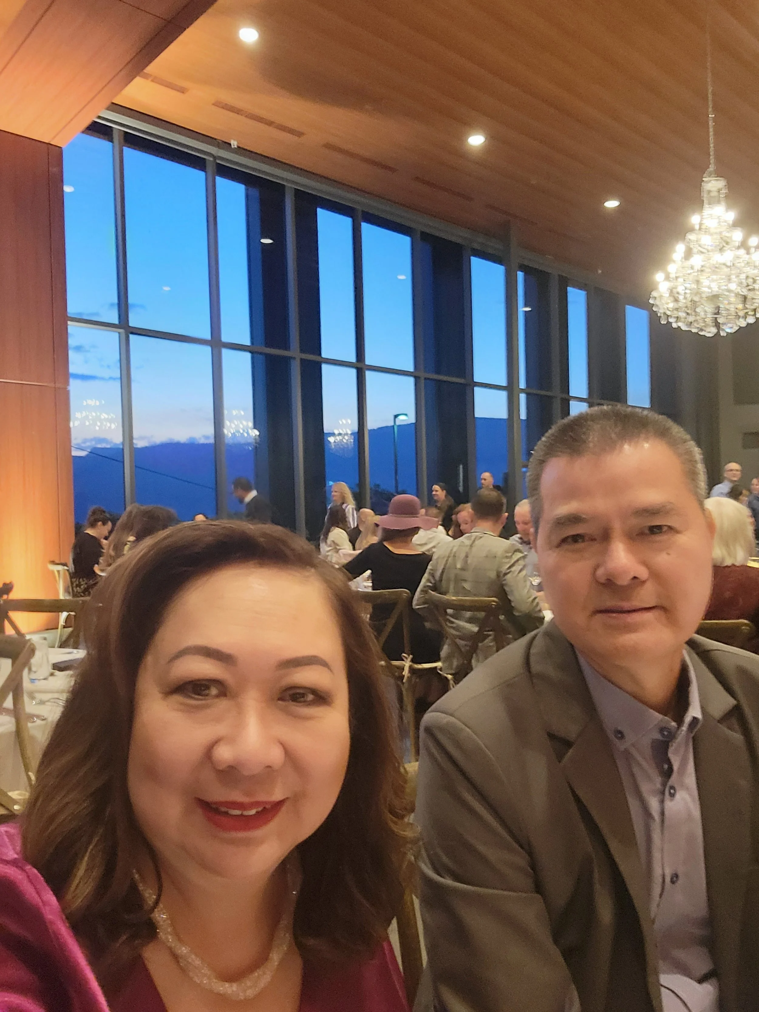 A woman and a man taking a selfie at a formal event in a spacious restaurant or banquet hall with large windows and a chandelier. The woman has brown hair and is wearing a pink outfit with a pearl necklace. The man has short gray hair and is wearing