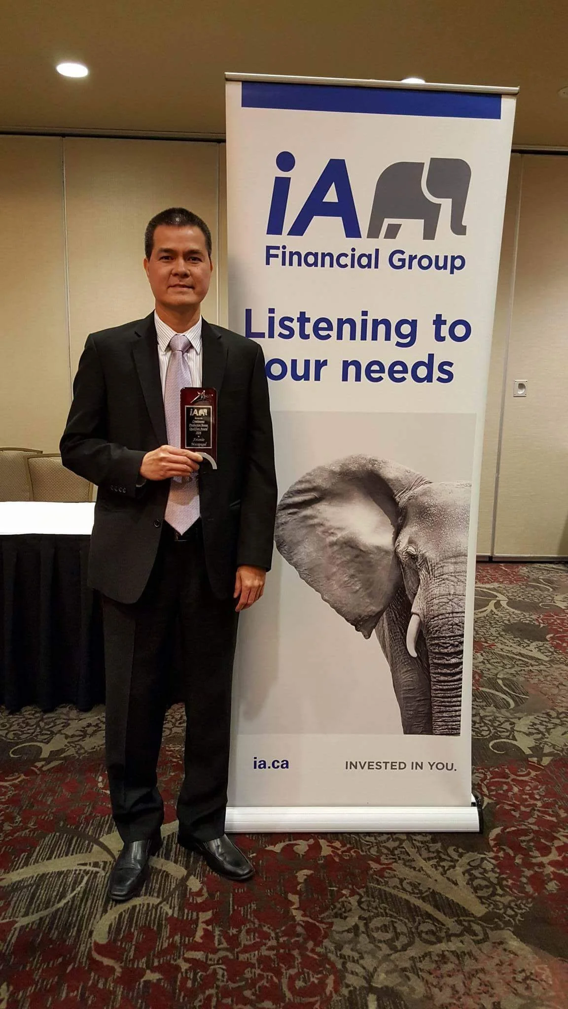A man in a black suit and tie standing beside a banner for iA Financial Group, which features an elephant image and the text "Listening to your needs."