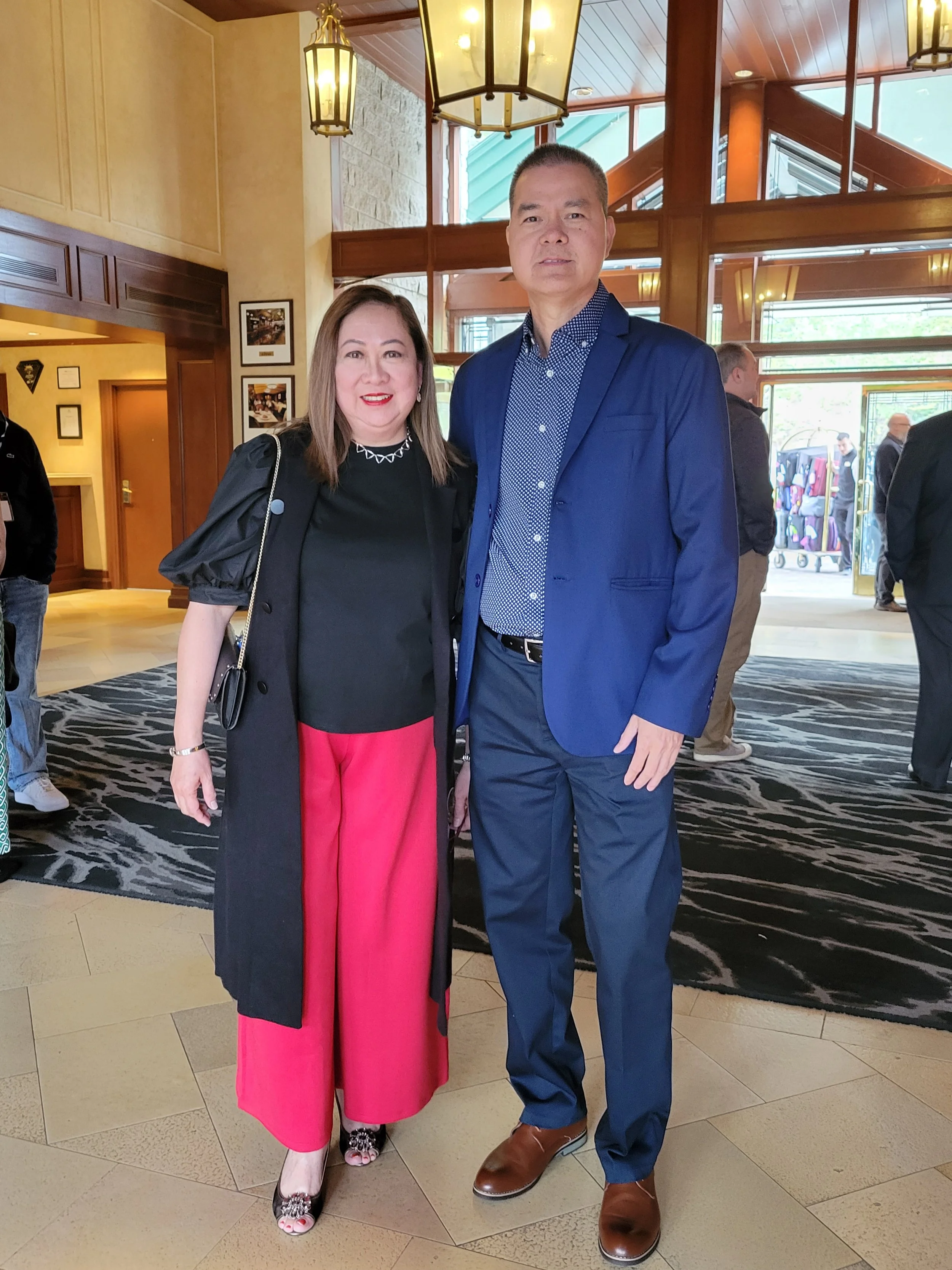 A woman and a man standing together indoors in a well-lit, elegant lobby. The woman is wearing a black top, red wide-leg pants, and high heels, with a black coat and a chain necklace. The man is wearing a blue suit with a patterned shirt and brown sh