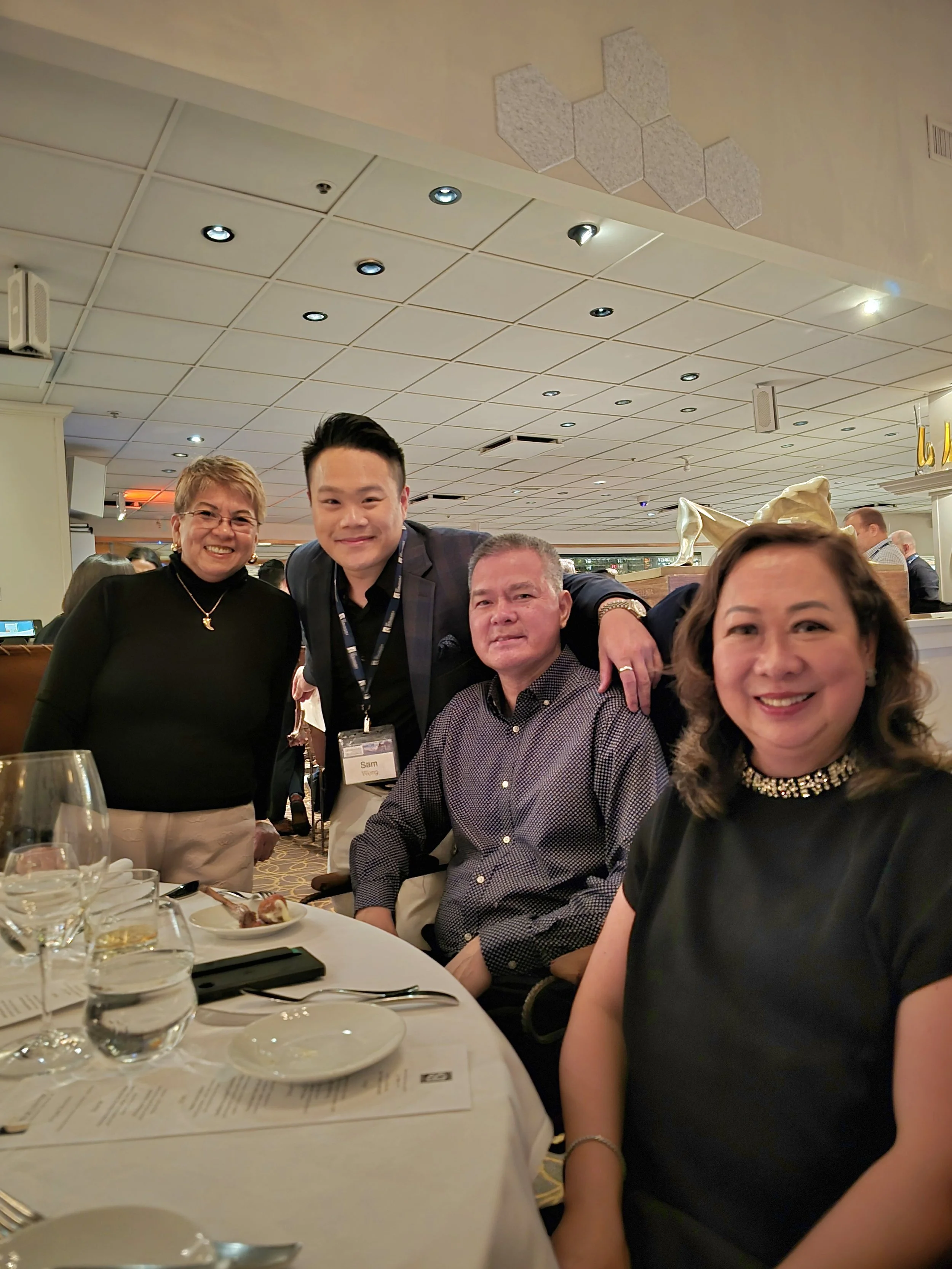 Group of four people smiling at a dinner event, some seated at a table with glasses, plates, and utensils, in a well-lit restaurant or banquet hall.