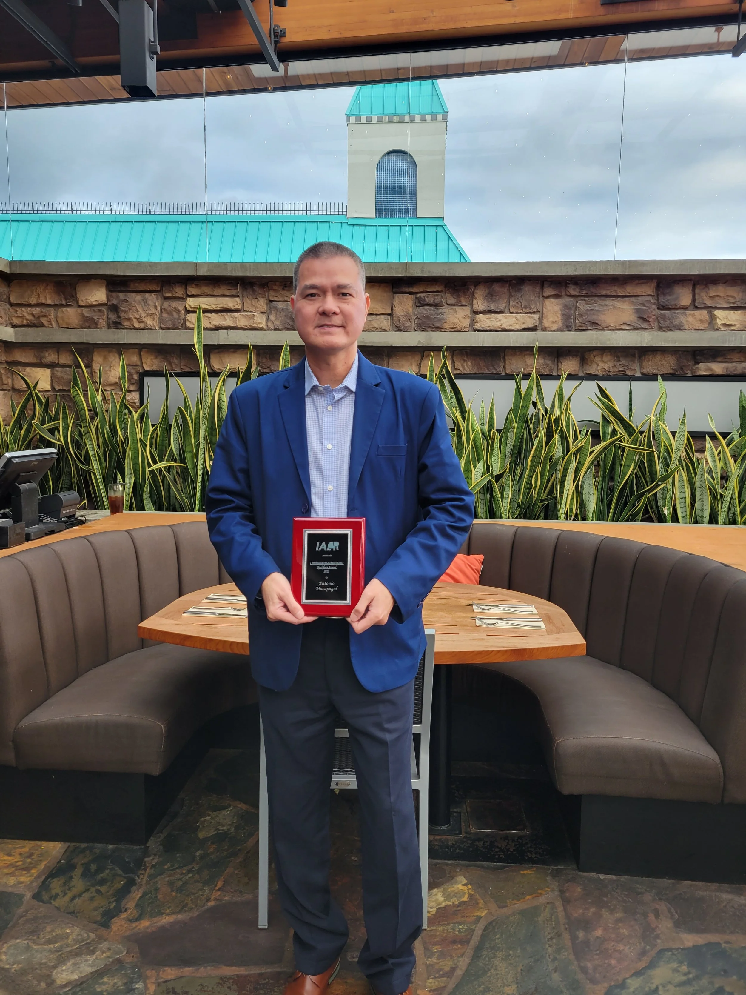A man in a blue blazer holding an award plaque in a restaurant with a curved bench, a wooden table, and plants, with a glass window showing a building with a clock tower in the background.