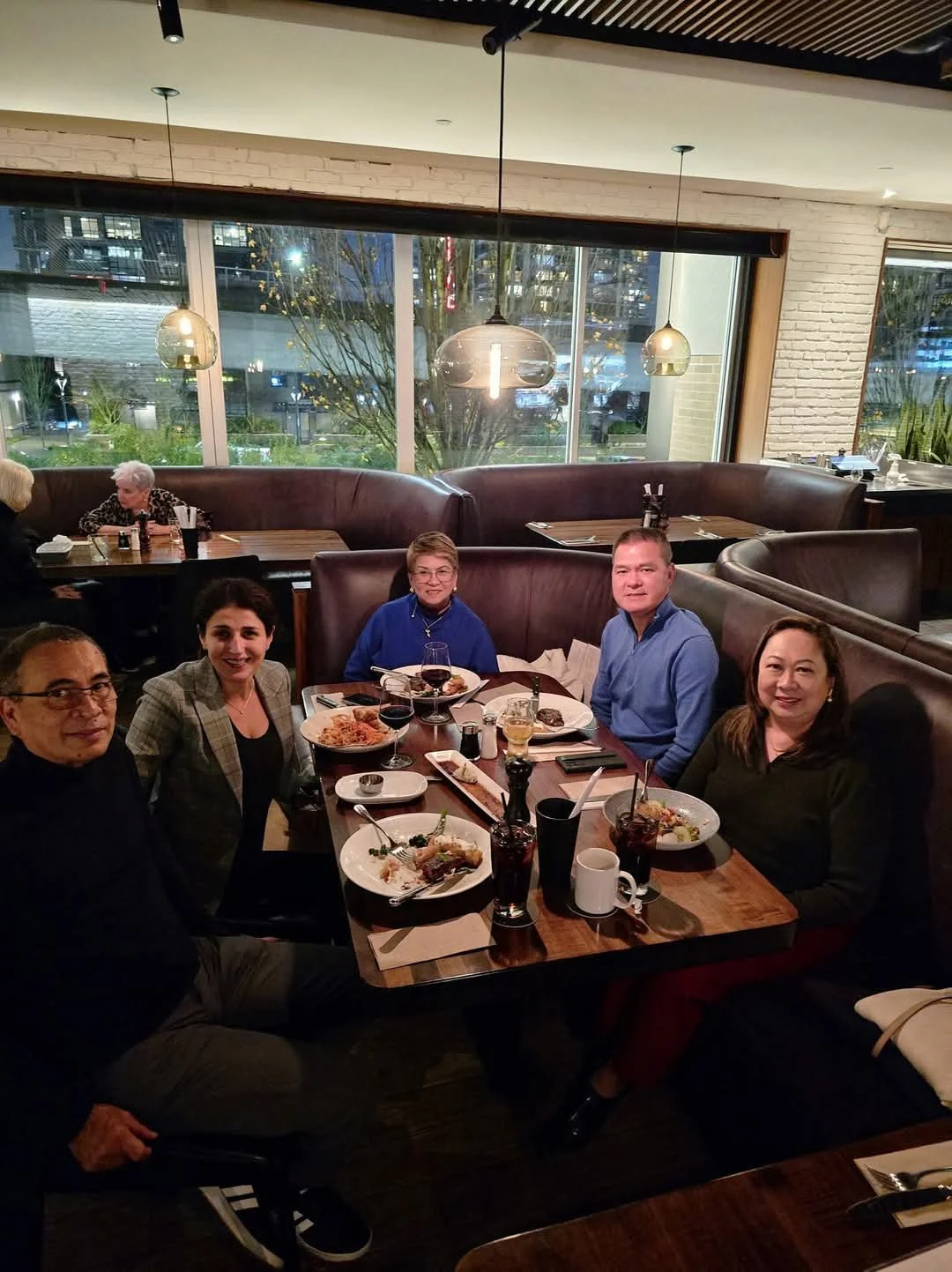 A group of five people sitting around a restaurant table with food and drinks, inside a modern restaurant with large windows and hanging pendant lights.