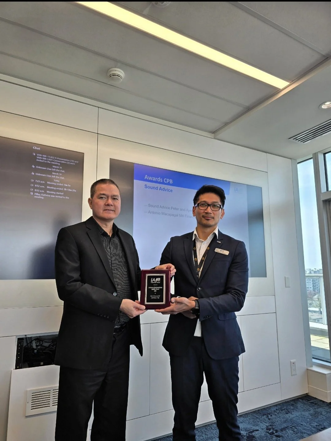 Two men in formal attire standing in a modern conference room, holding a plaque or award together. One man has gray hair and a black suit, the other has dark hair, glasses, and a navy blue suit. A large screen behind them displays a presentation slid