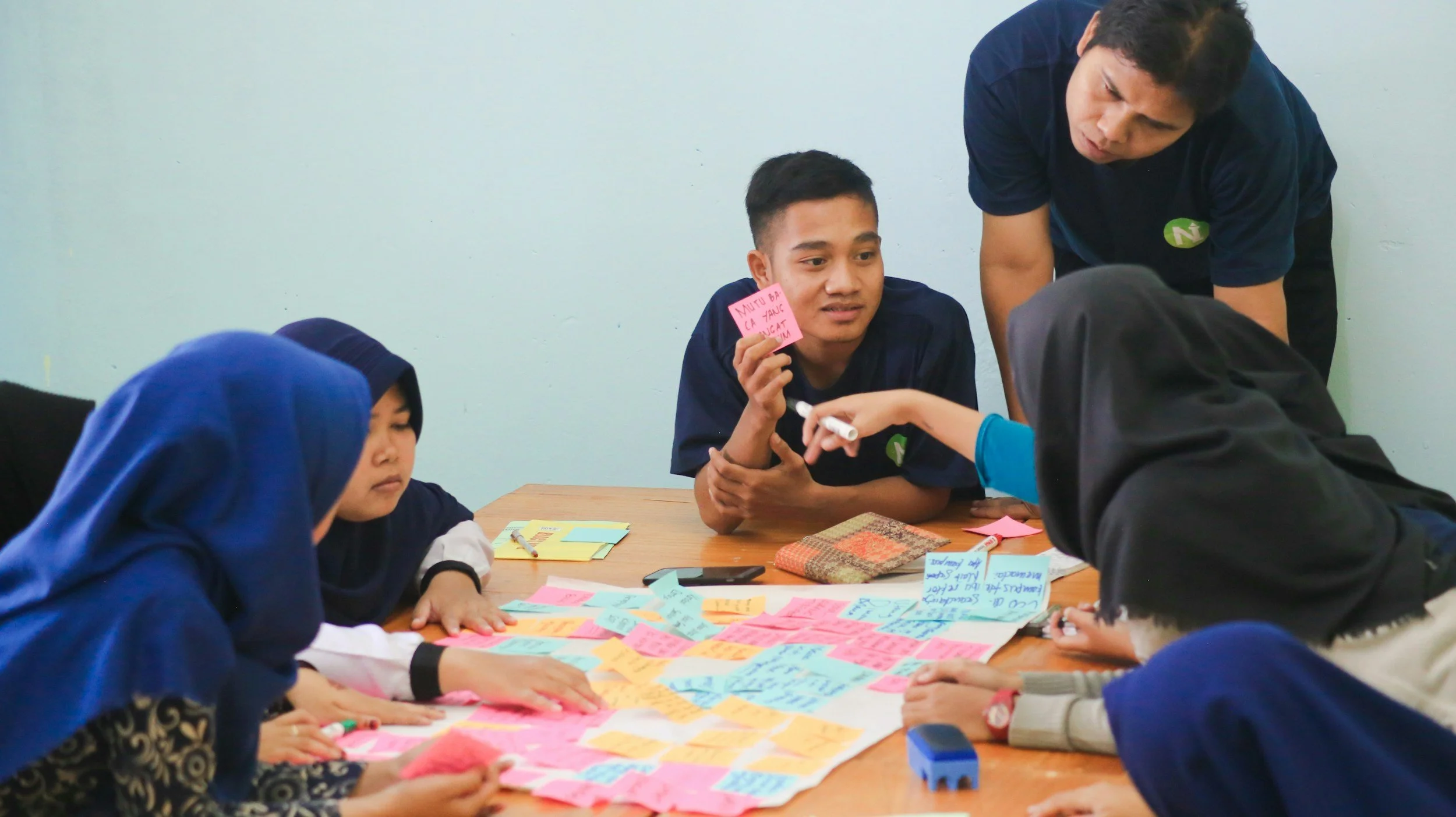 A group of young students, some wearing hijabs, sitting around a table with colorful sticky notes and papers, engaged in a collaborative activity. One student holds a pink note, and a teacher is assisting the students.