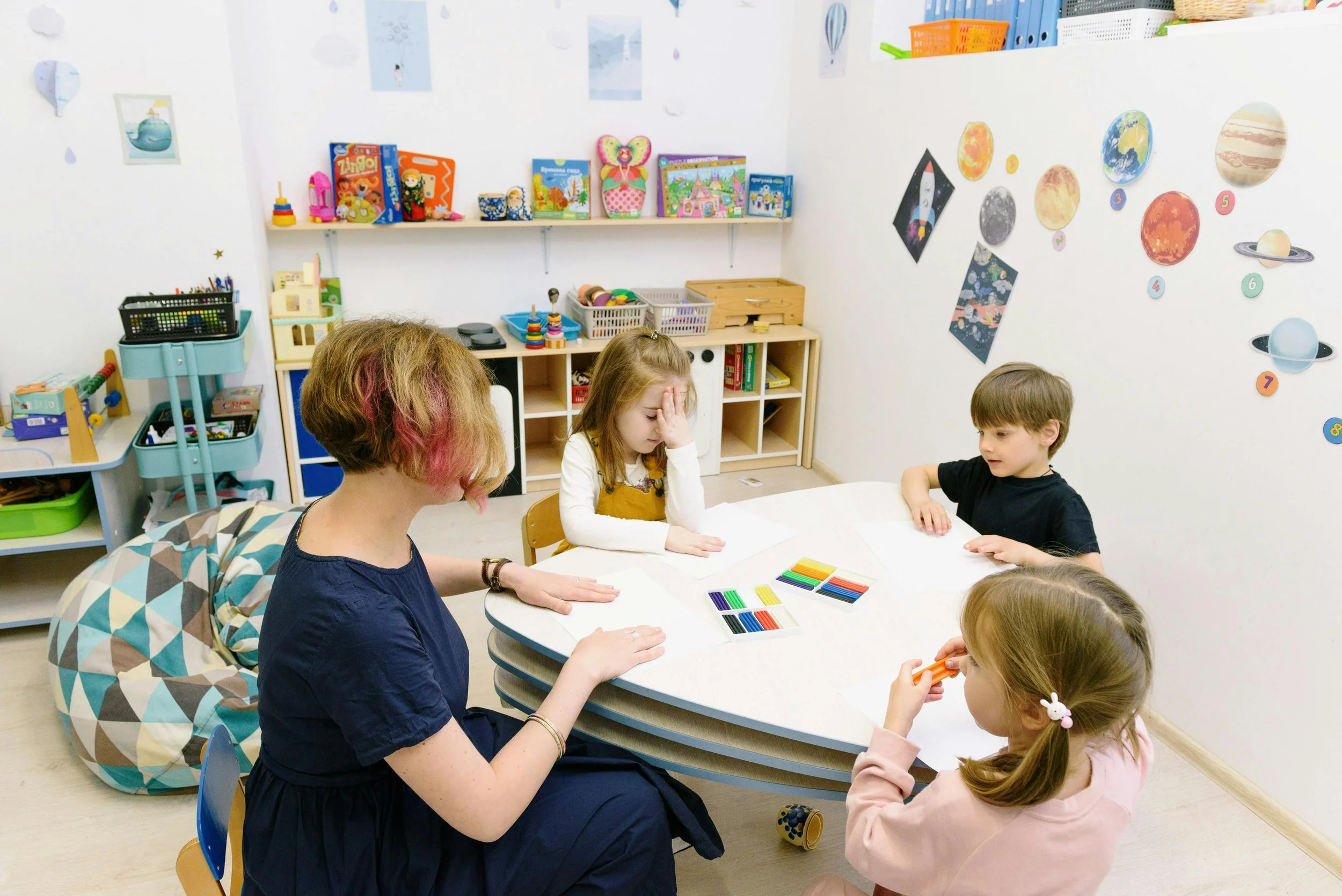 A woman and four children sitting at a table in a classroom. The woman appears to be overseeing or assisting the children with an activity involving colorful modeling clay or playdough. The classroom has space-themed decorations on the wall, including images of planets and a rocket, and shelves filled with toys, books, and educational materials.