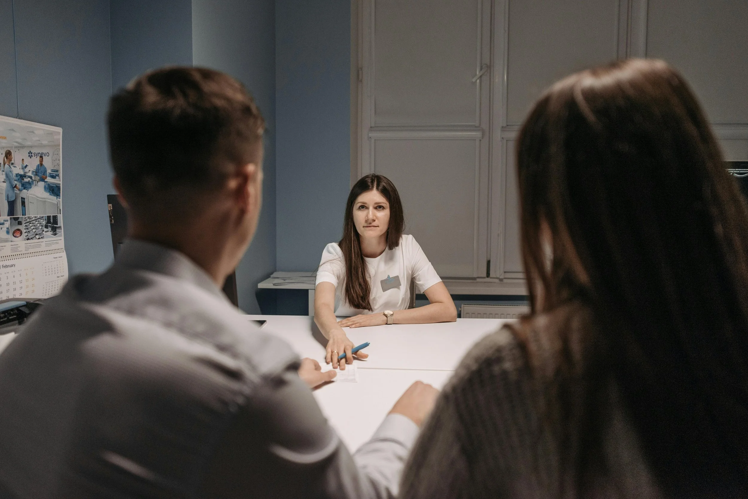 A woman with long brown hair sitting at a table during a meeting or interview, facing forward with two people whose backs are to the camera, in a professional office setting.
