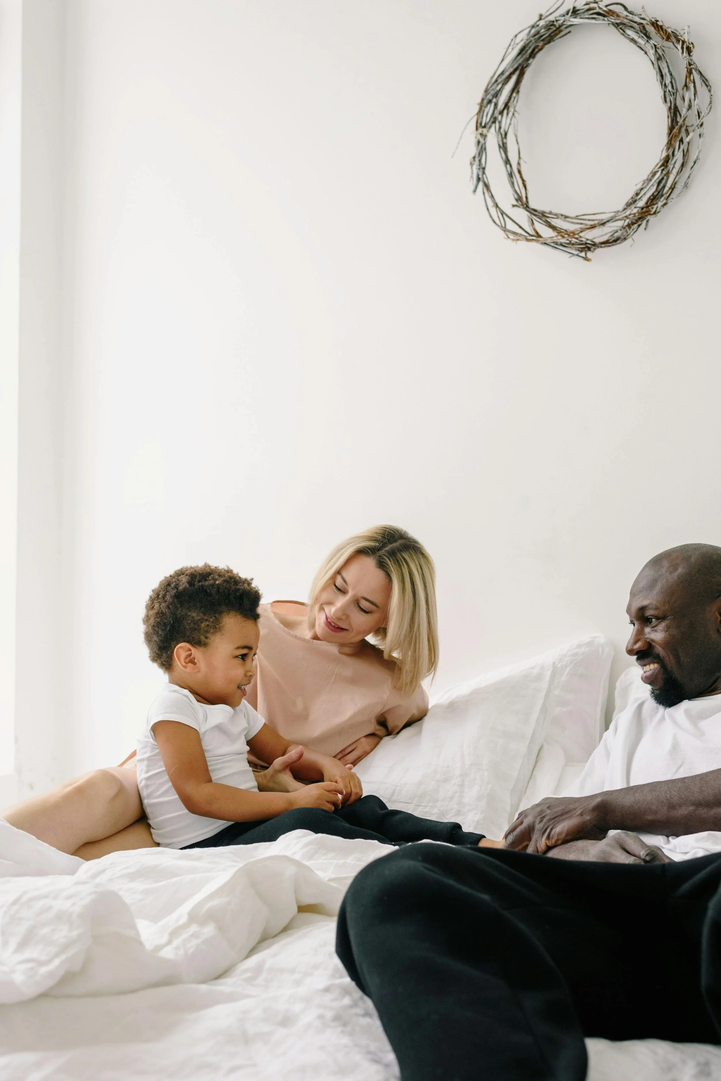 A family of four sitting on a bed, smiling and talking together in a bright, minimal bedroom with a wreath hanging on the white wall.