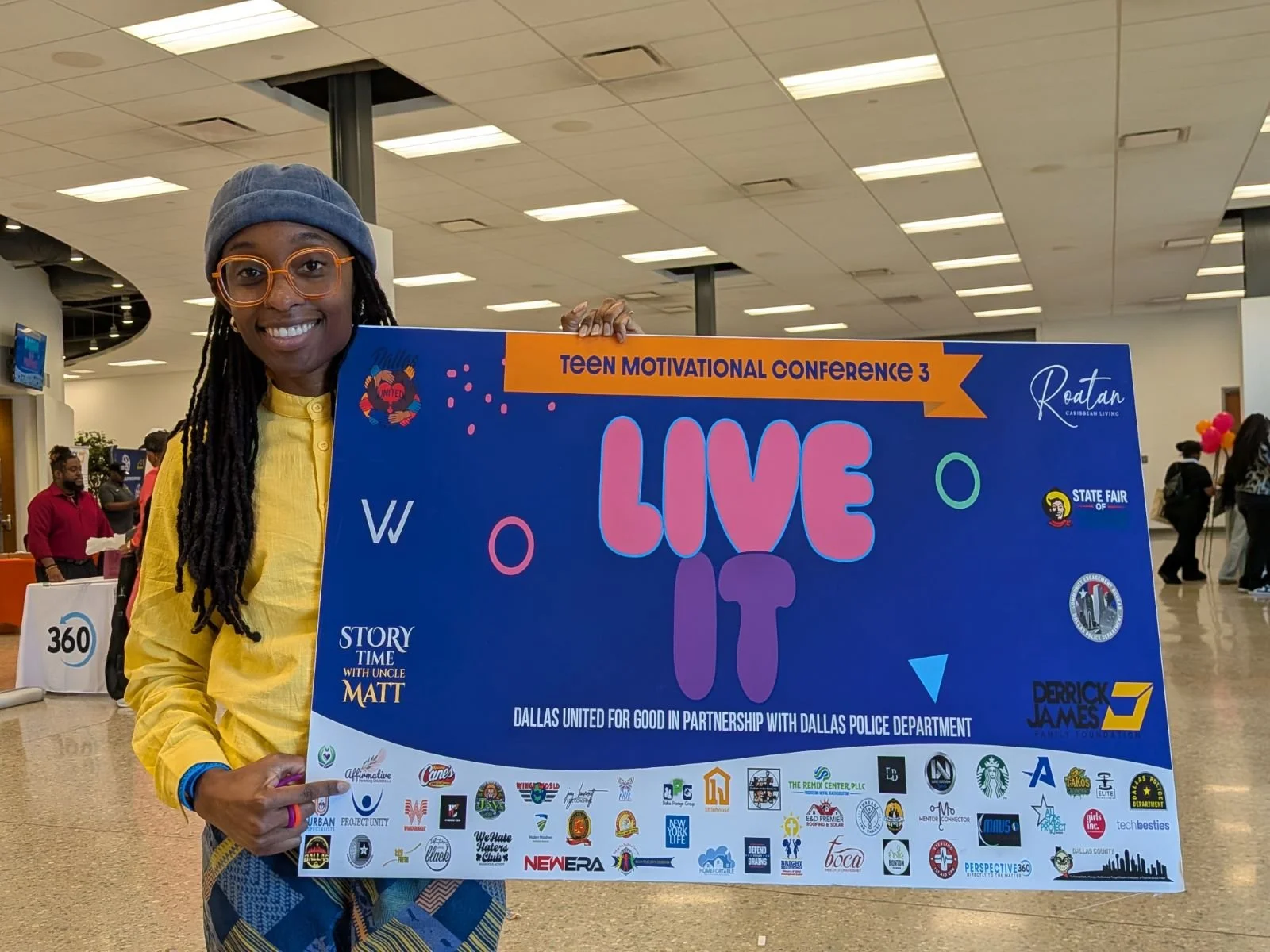 A person smiling and holding a large colorful sign at an indoor event, with other people in the background.