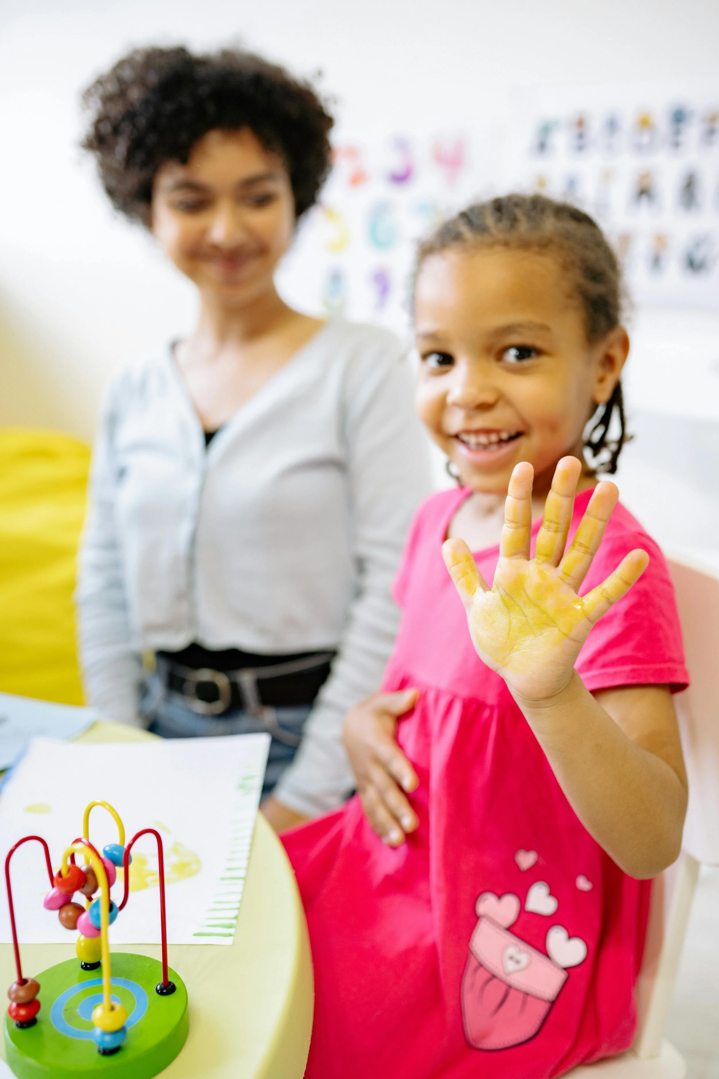 A young girl with braided hair and a pink dress showing a yellow painted hand, smiling and giving a peace sign. Behind her is a woman with curly hair, blurred in the background. In the foreground, there is a colorful bead maze toy on a yellow table.