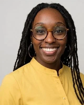 A woman with long, dark, styled hair wearing glasses with round frames and a yellow top, smiling at the camera against a neutral background.