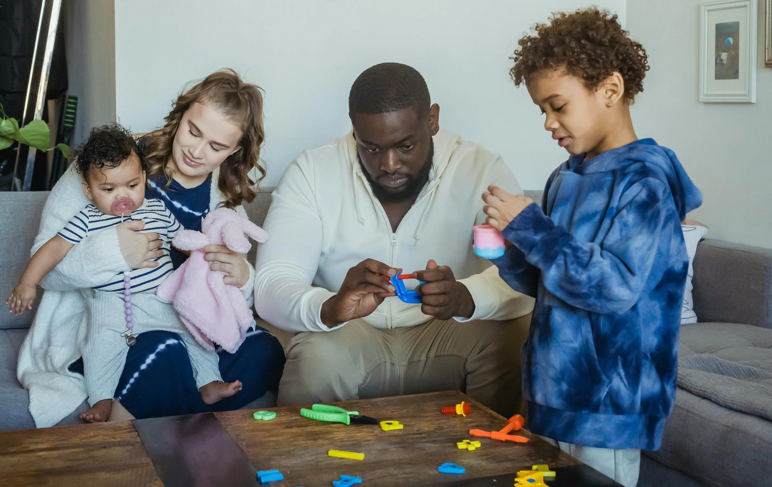 A family of five, including a toddler, two children, and two adults, gathered around a wooden table playing with colorful plastic letters and toys in a living room.