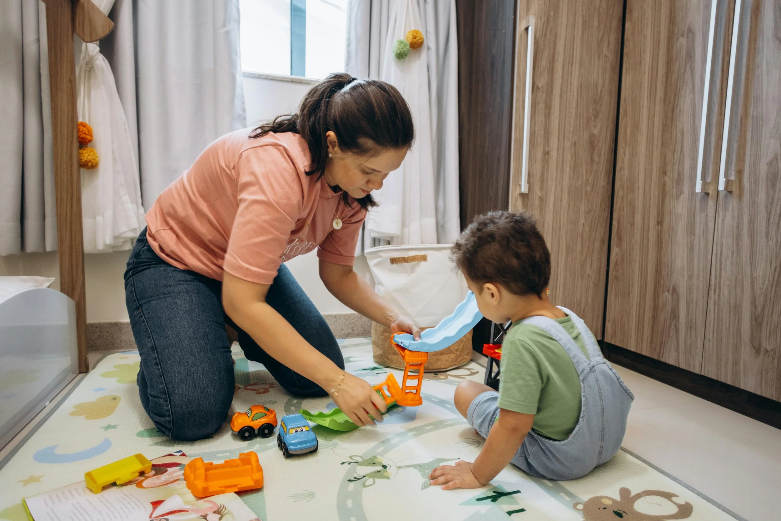 A woman and a young boy playing with toy cars in a room with wooden cabinets and a large window.