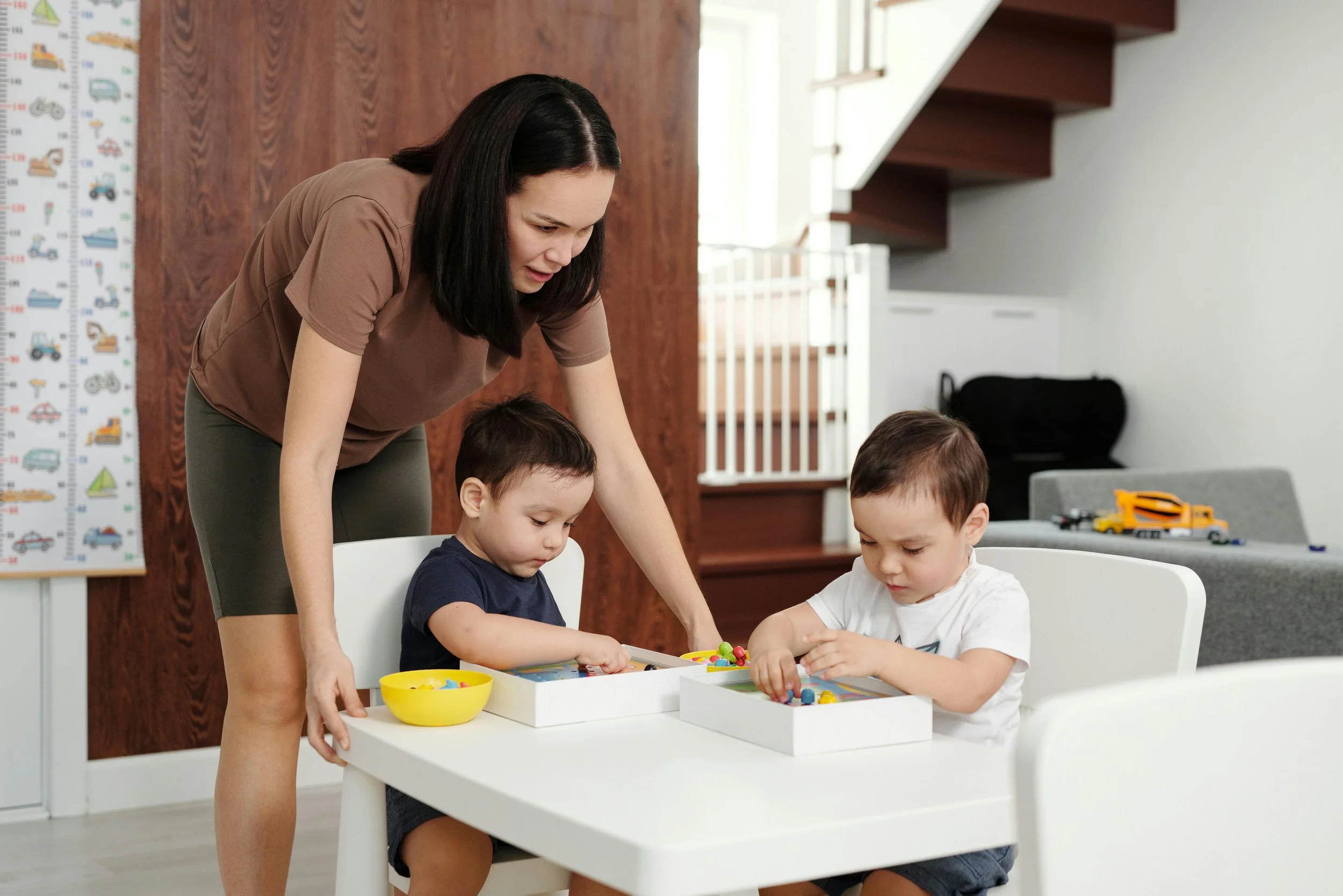 A woman helping two young boys with a board game at a white table in a bright room.