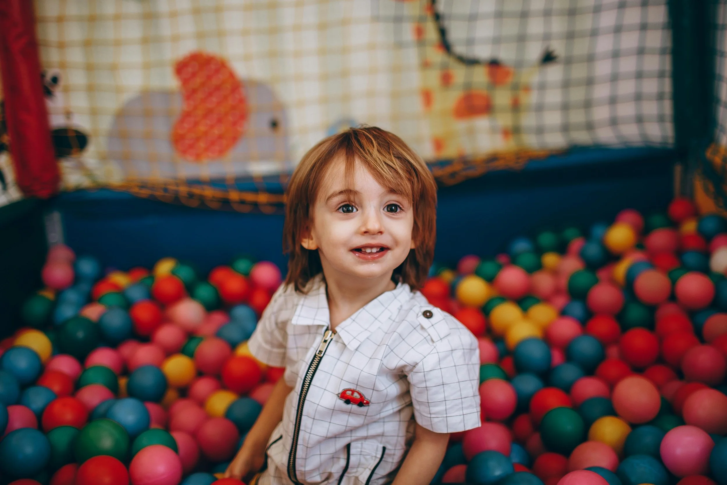 A young boy with red hair and a checkered shirt playing in a colorful ball pit inside an indoor play area.