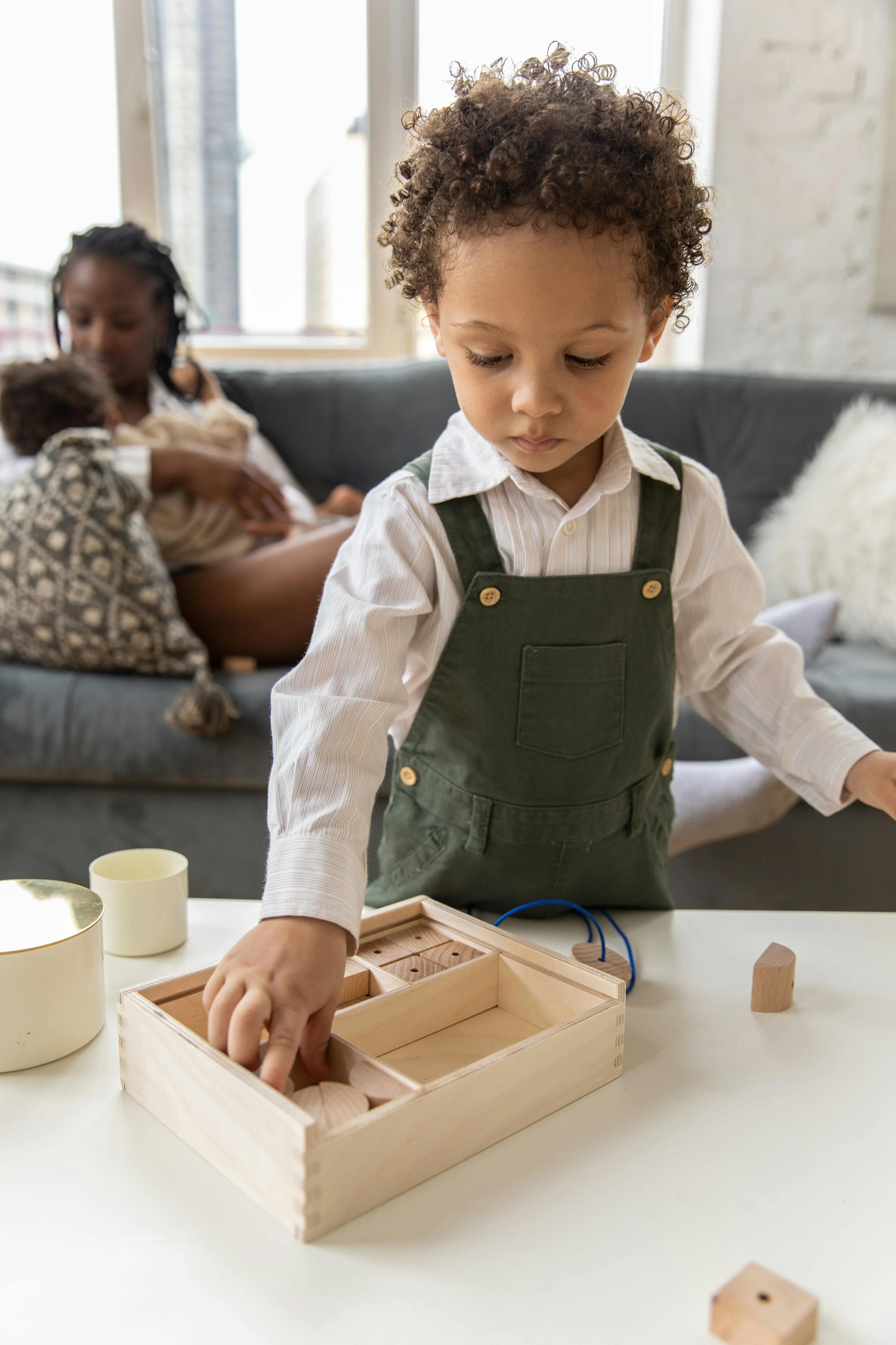 A young boy with curly hair wearing a white shirt and green overalls is playing with wooden blocks and toys at a table. In the background, a woman with braided hair is holding a child, sitting on a gray couch near a window in a bright room.