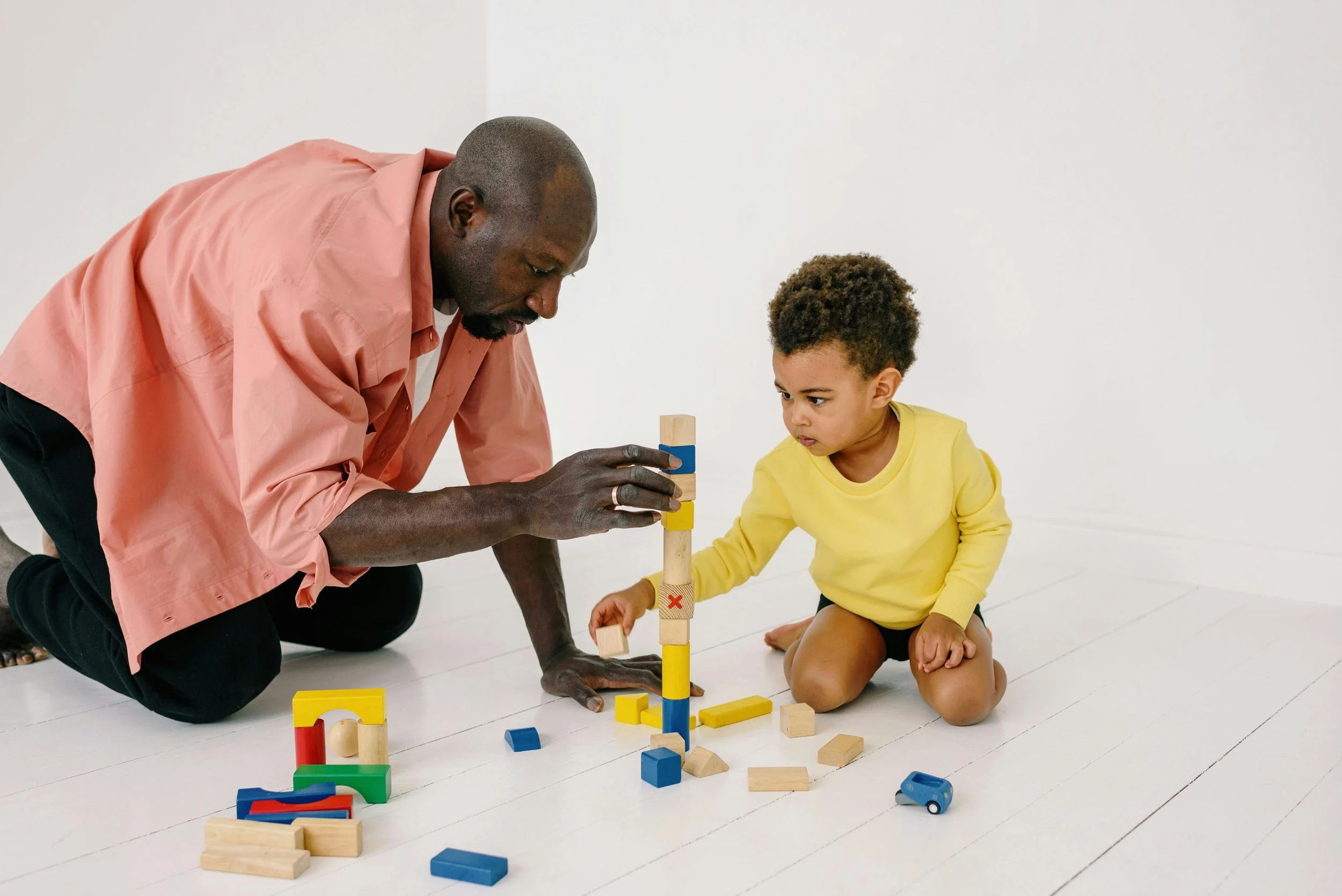 An adult man and a young girl engaging in a creative activity with wooden building blocks and toy vehicles on a white floor in a minimalistic setting.