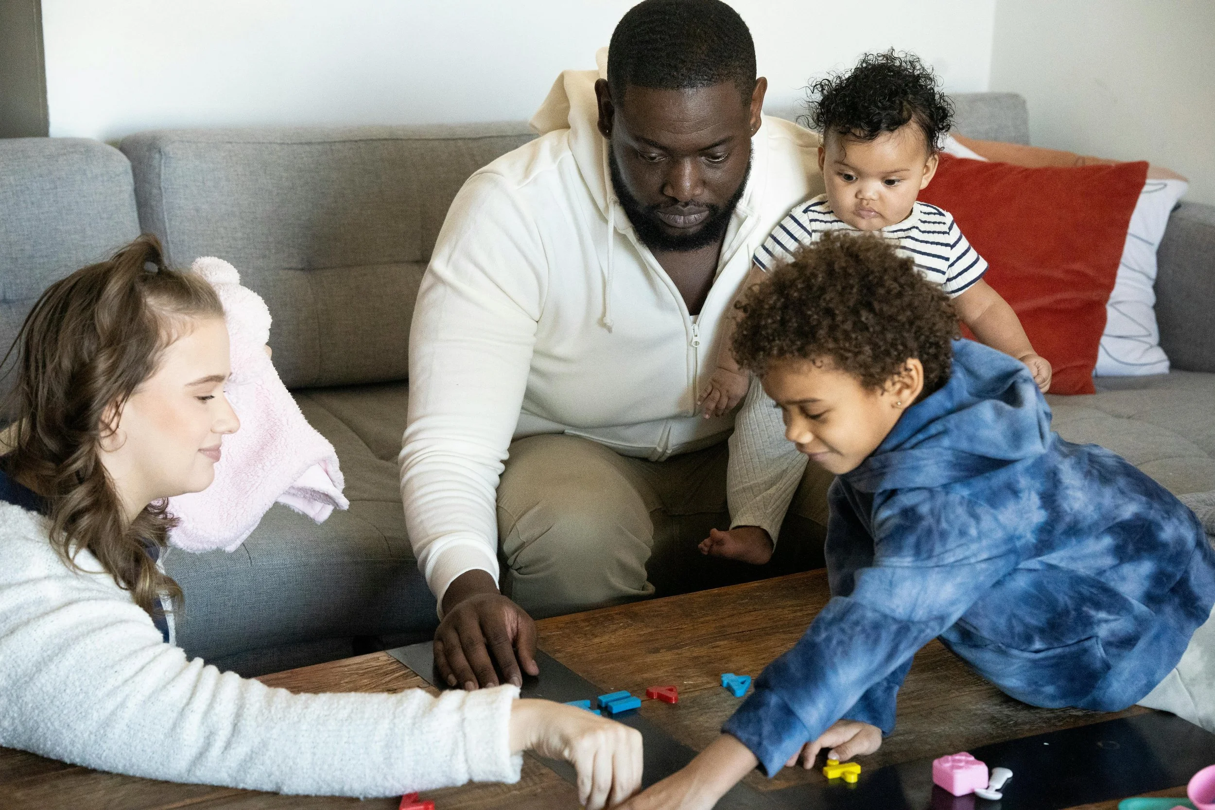A man and four children sitting on a couch, playing a game with colorful pieces on a wooden table in a living room.