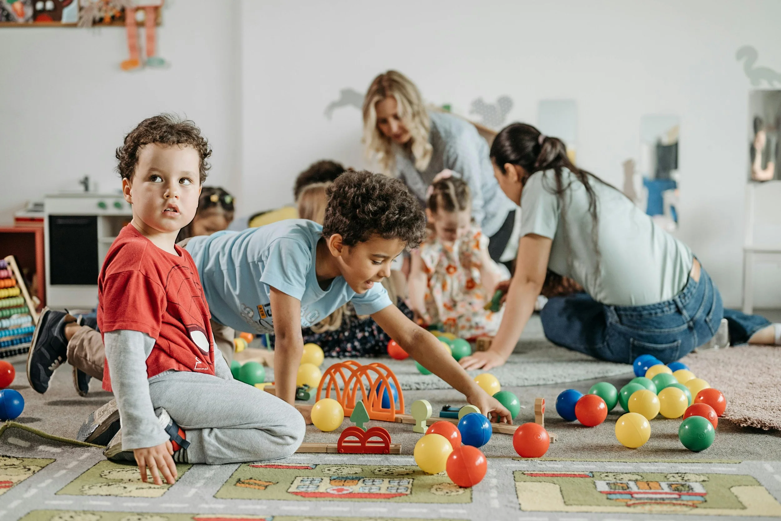 Children playing with colorful balls and toys in a classroom while adults supervise, on a patterned carpet with toys and decorations on walls.