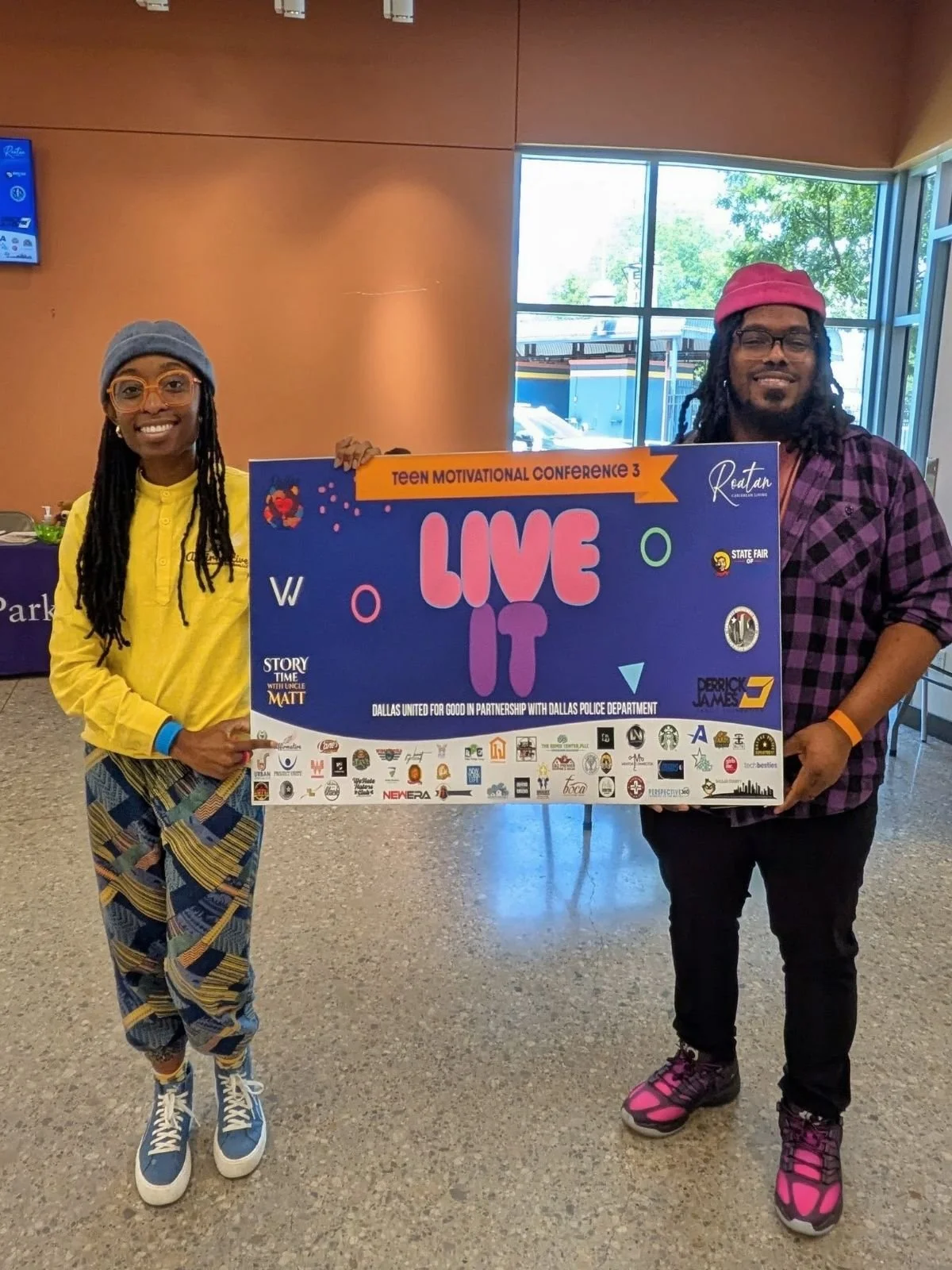 Two people holding a colorful sign at an indoor event. The sign reads 'LIVE IT' with various logos and text underneath, including 'Teen Motivational Conference 3' and 'Dallas United for Good in partnership with Dallas Police Department.' The woman on the left wears a gray beanie, glasses, a yellow shirt, patterned pants, and sneakers. The man on the right wears a pink beanie, glasses, a purple checkered shirt, black pants, and pink sneakers.