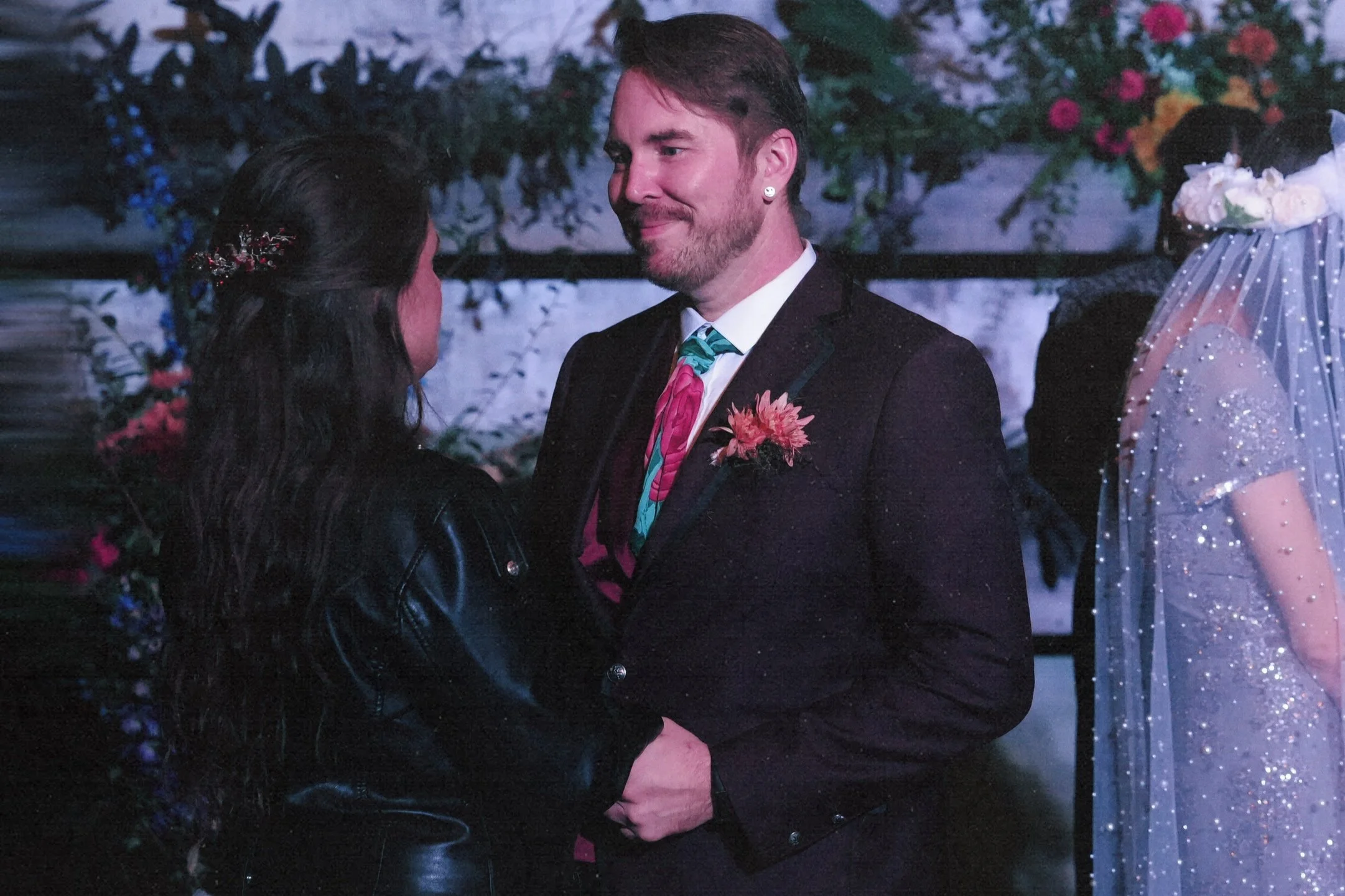 A man in a suit with a pink flower boutonniere holds hands with a woman with dark hair and a floral hair accessory, at a wedding reception or similar event.