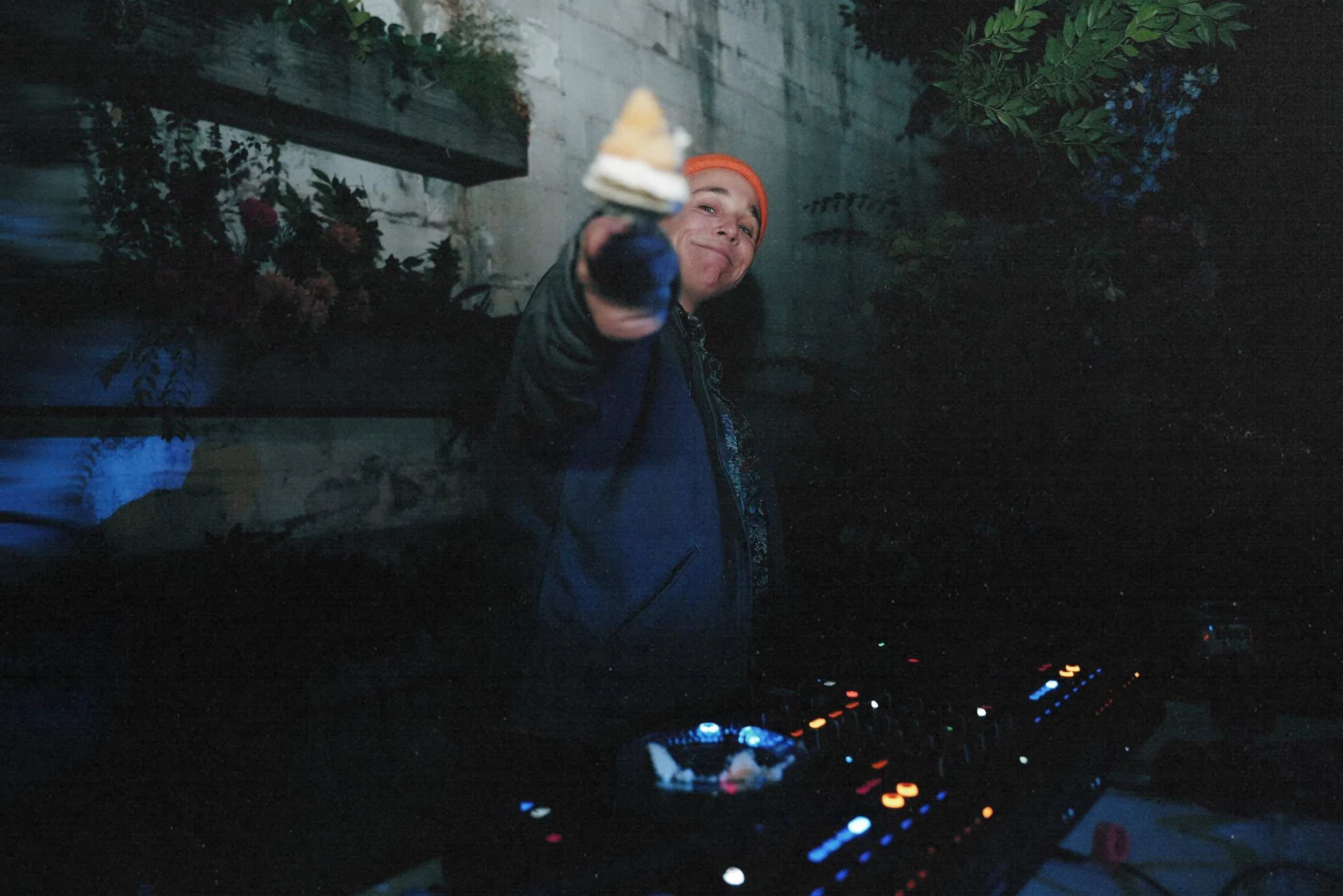 A person wearing an orange beanie and a dark jacket, smiling, holding a slice of cake towards the camera, with DJ equipment in front of them, in an outdoor setting at night.